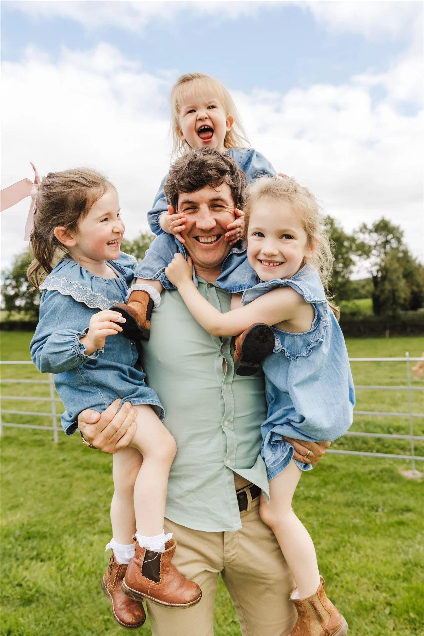 Dad carrying three young girls, all smiling and laughing outdoors at home in garden with trees in the background.