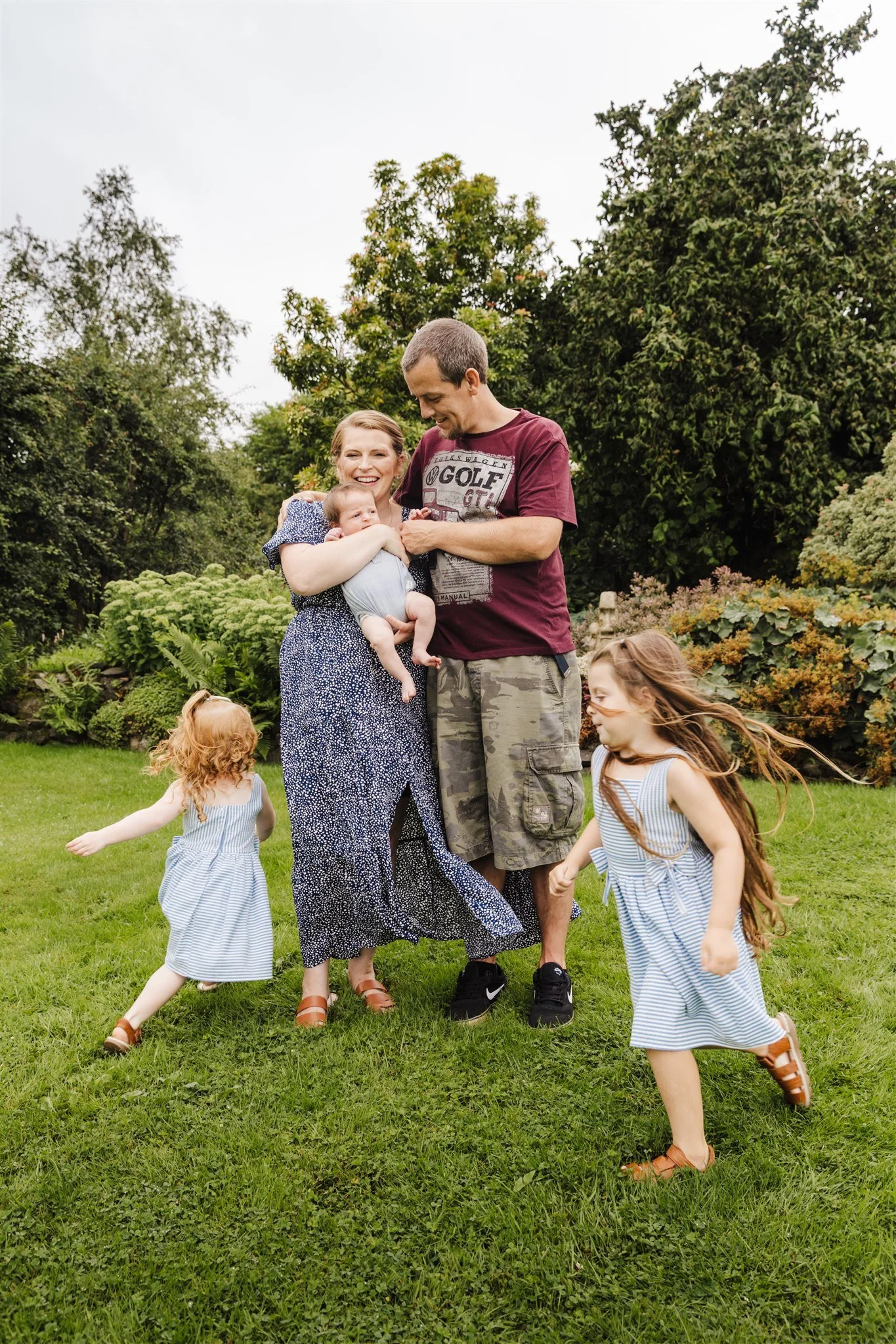 A woman holding a baby and a man, with two young girls playing around them on a grassy lawn in a garden.