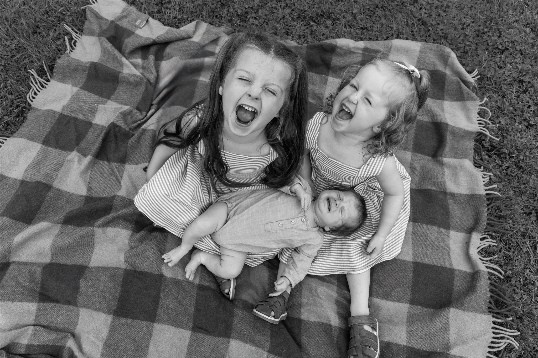 Three young children, two girls and one baby, lying on a checkered blanket outdoors, laughing and playing. Sibling photos outside