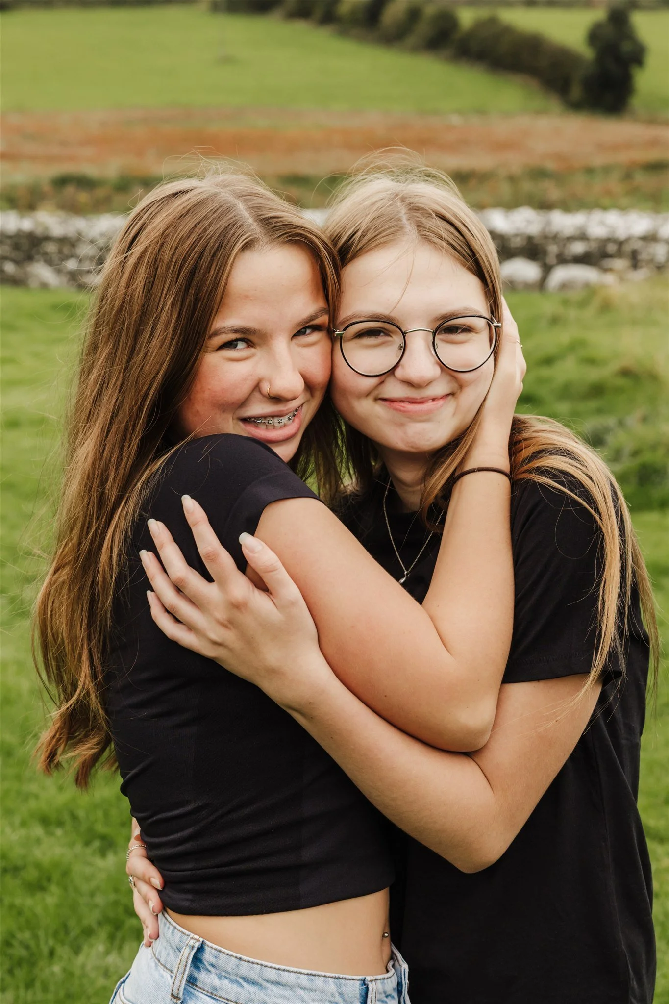 Two sisters hugging outdoors in a grassy area with a small river and trees in the background. Sisters Portrait photography