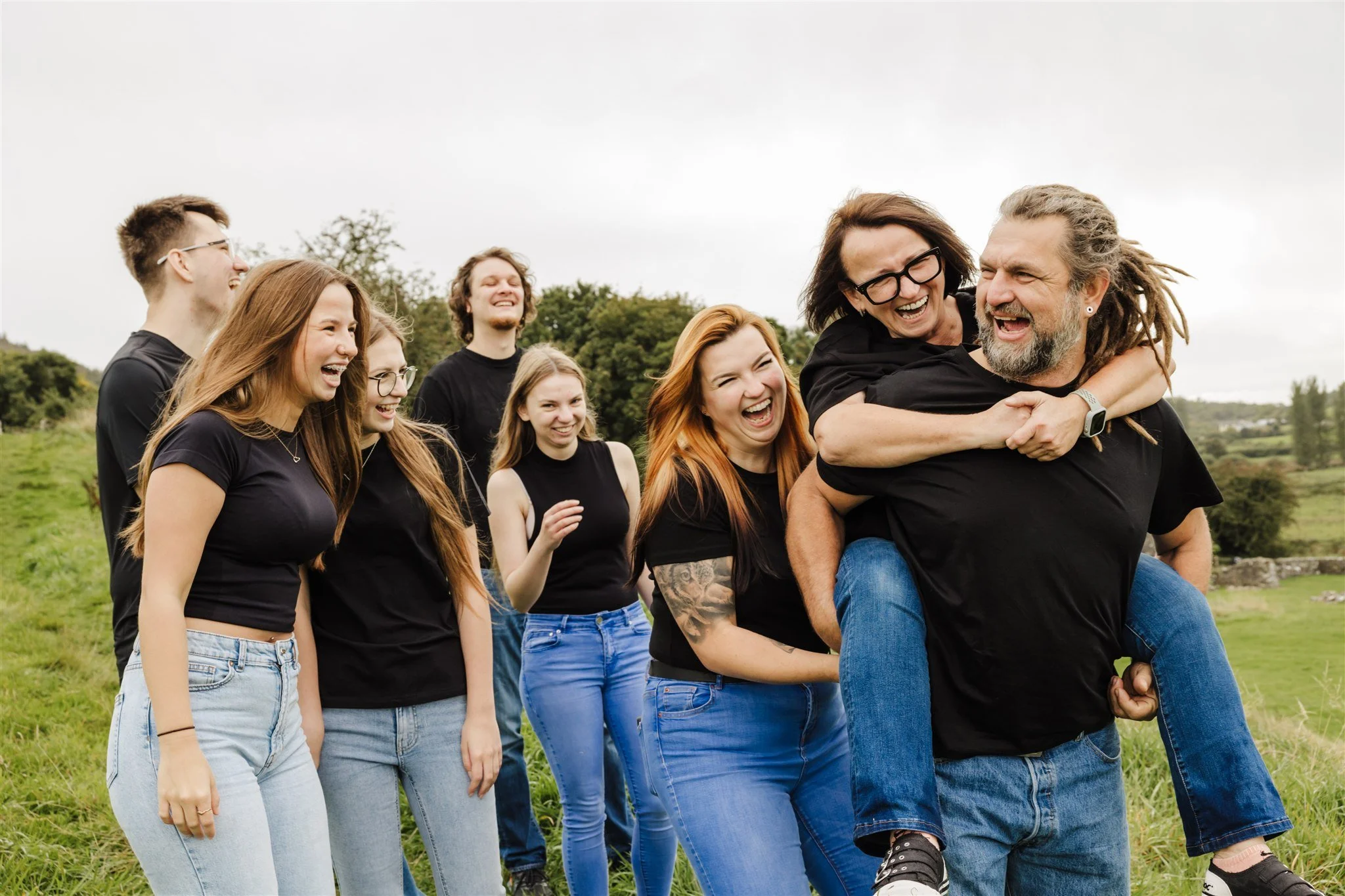Group of seven friends outdoors in a grassy area, laughing and enjoying each other's company. One person is piggybacking on another, with everyone smiling and having fun.