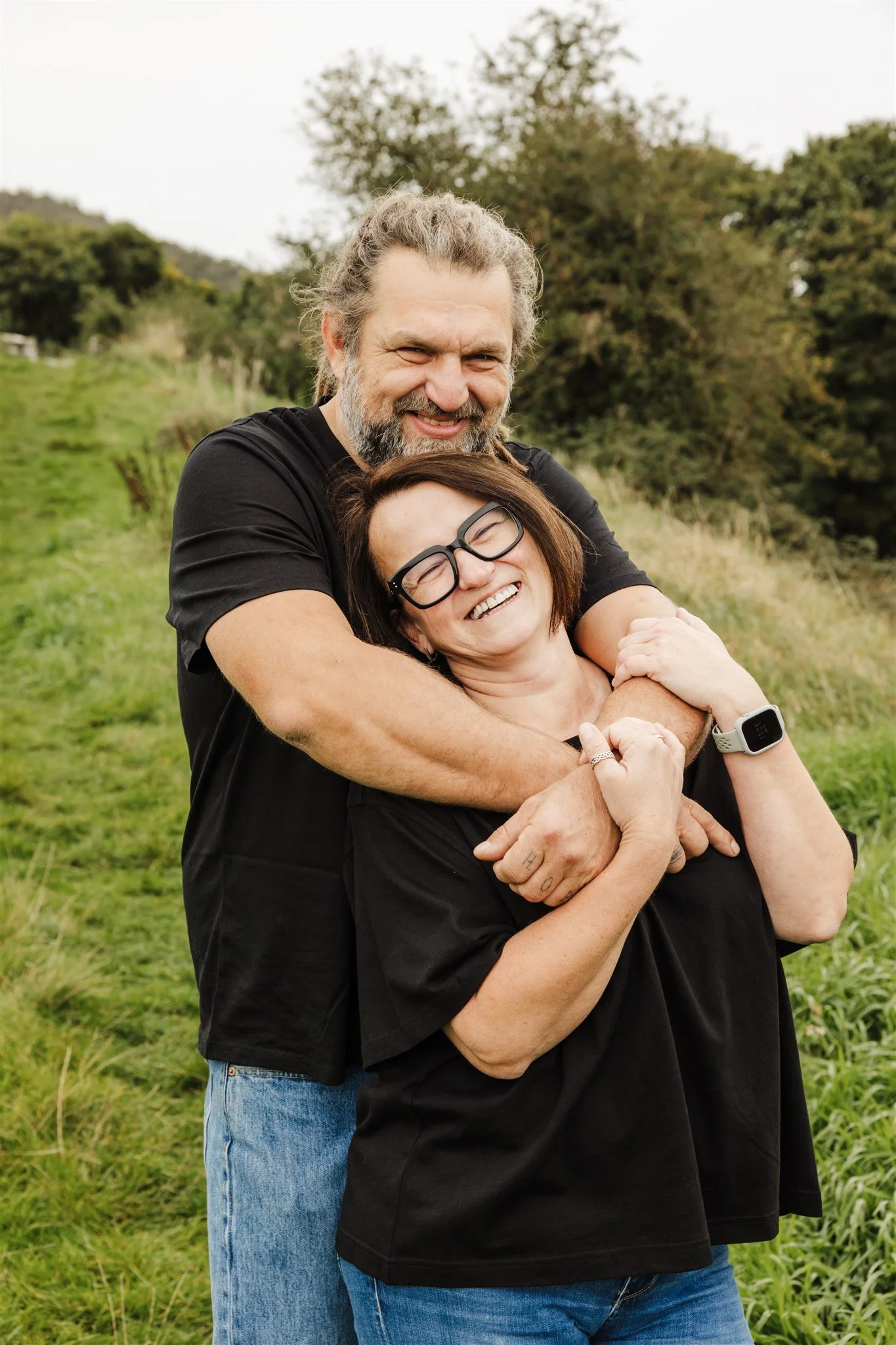 A happy man with a beard hugging a smiling woman with glasses outdoors in a grassy field with trees in the background.