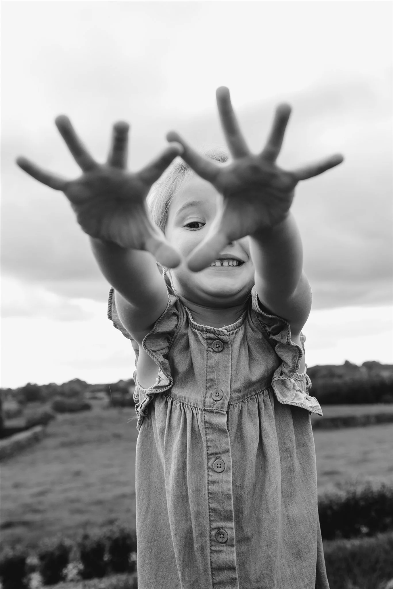 A young girl in a sleeveless dress smiling and reaching forward with open hands, outdoors with a cloudy sky and landscape in the background.