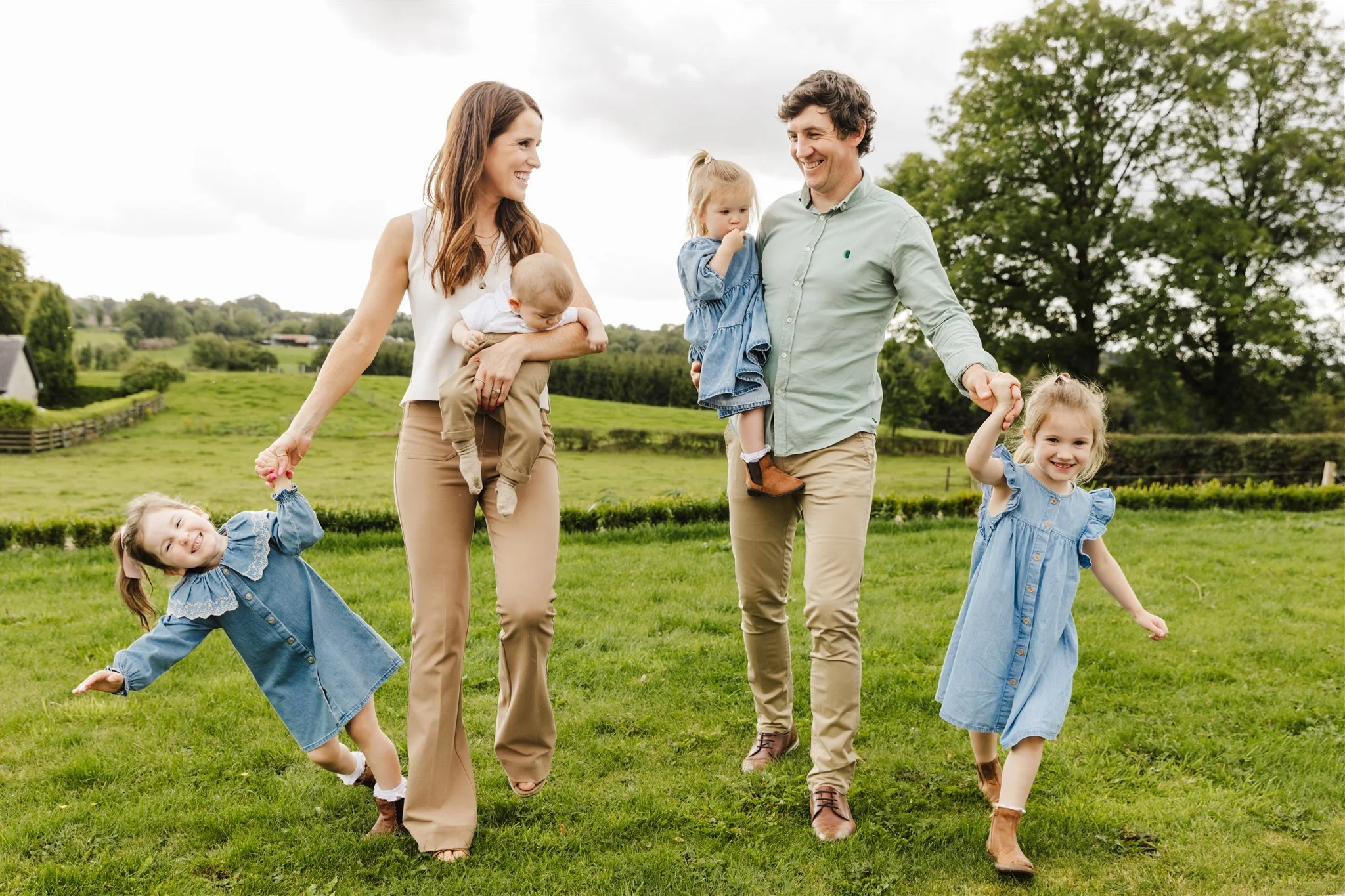 Family of six enjoying a day outdoors on a grassy field with trees in the background. The mother, father, and four children are smiling, holding hands, and walking together.