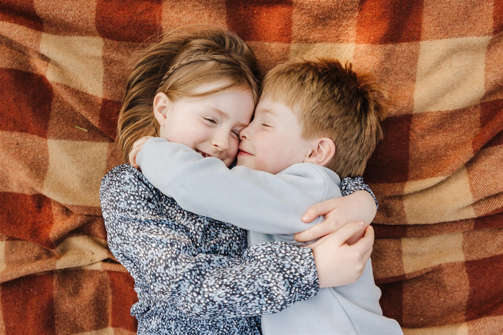 Two children, a girl and a boy, hugging and smiling while lying on a checkered blanket.