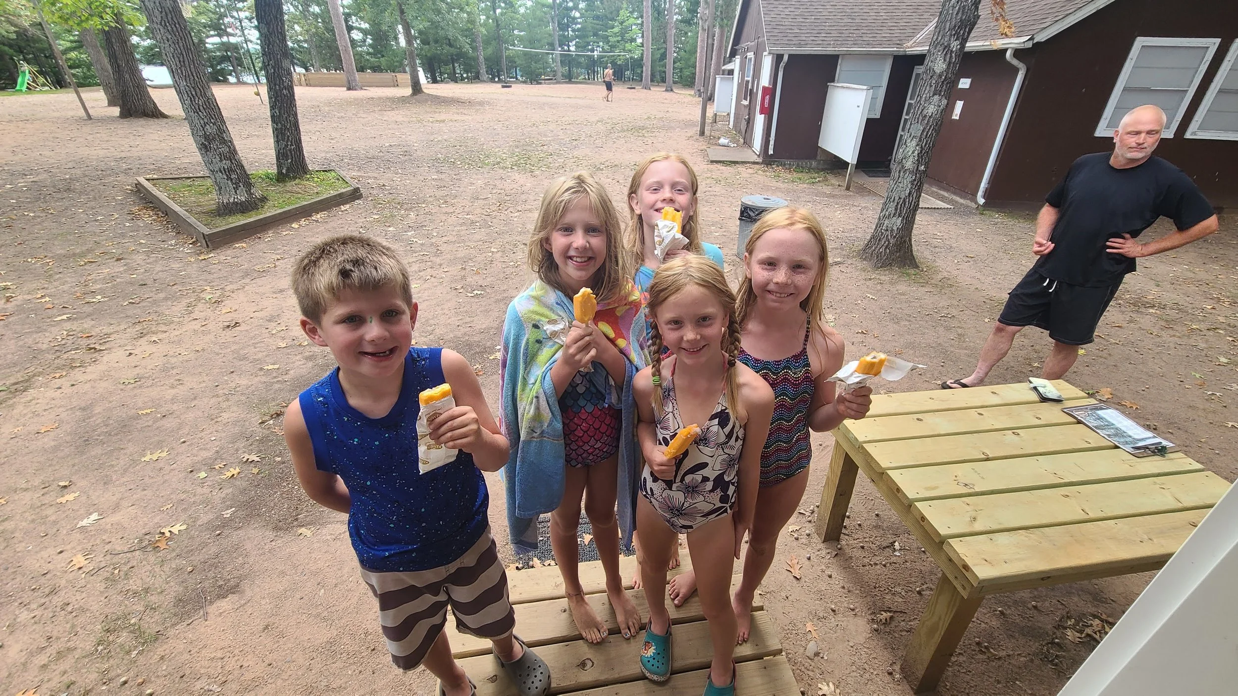 Six children in swimsuits smiling and holding popsicles at an outdoor park with trees and a building in the background. A man stands near a wooden picnic table on the right.