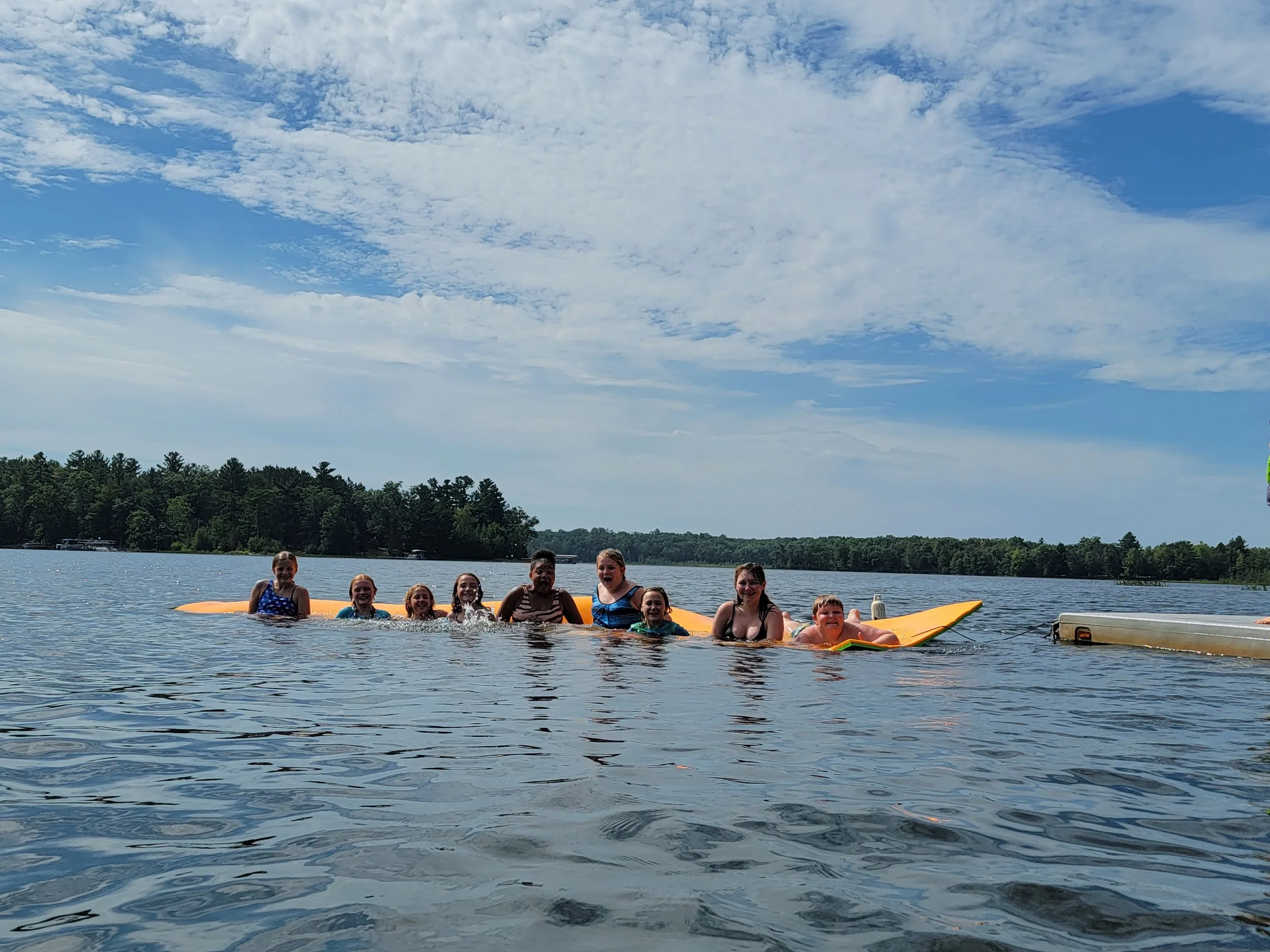 Group of children on a yellow boat in a lake, smiling and enjoying a sunny day, with trees and blue sky with clouds in the background.