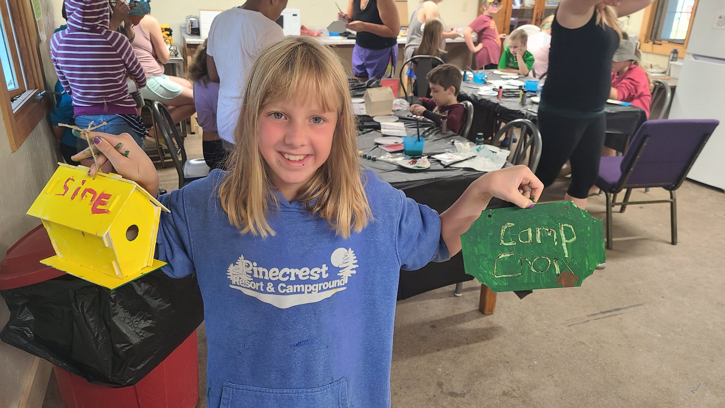 A young girl smiling and holding two painted birdhouses, one yellow and one green, in a room with other children and adults engaged in arts and crafts.