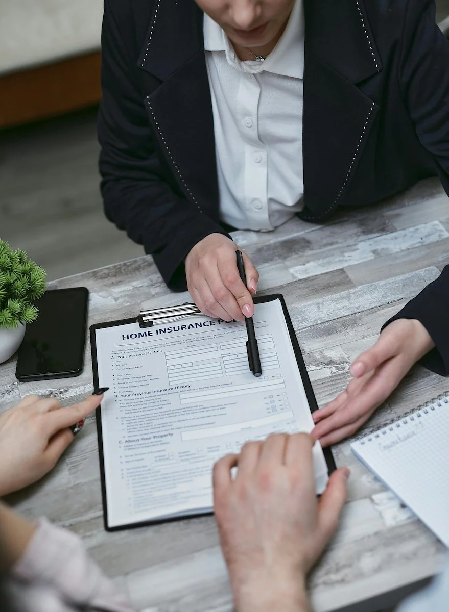 Person filling out a home insurance form on a clipboard while others are discussing the form. There is a smartphone, a notepad, and a small potted plant on the table.