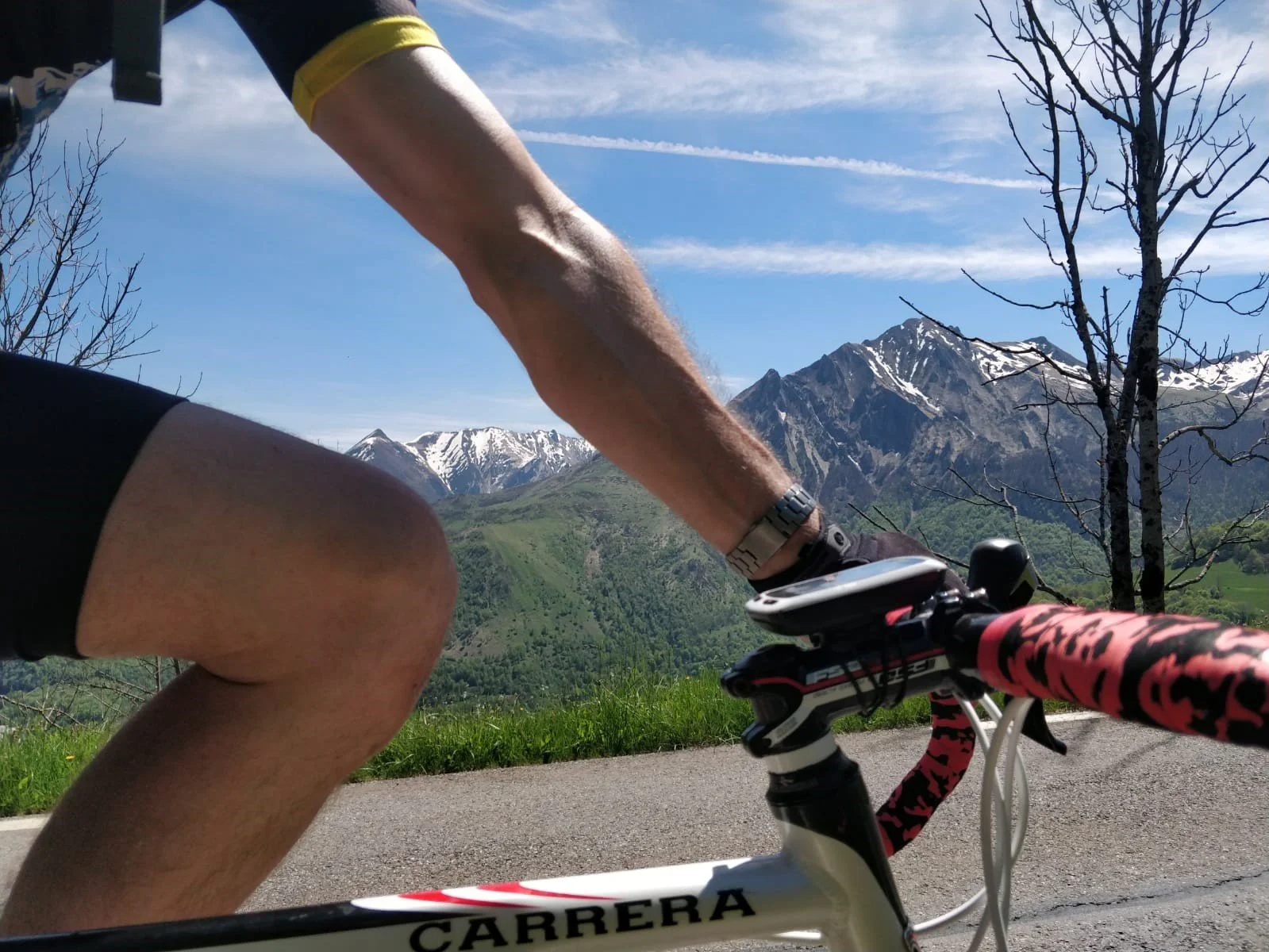 A person riding a bike through a mountainous landscape with snow-capped peaks and a clear blue sky, seen from a low angle focusing on the cyclist's arm and the bike's handlebars.