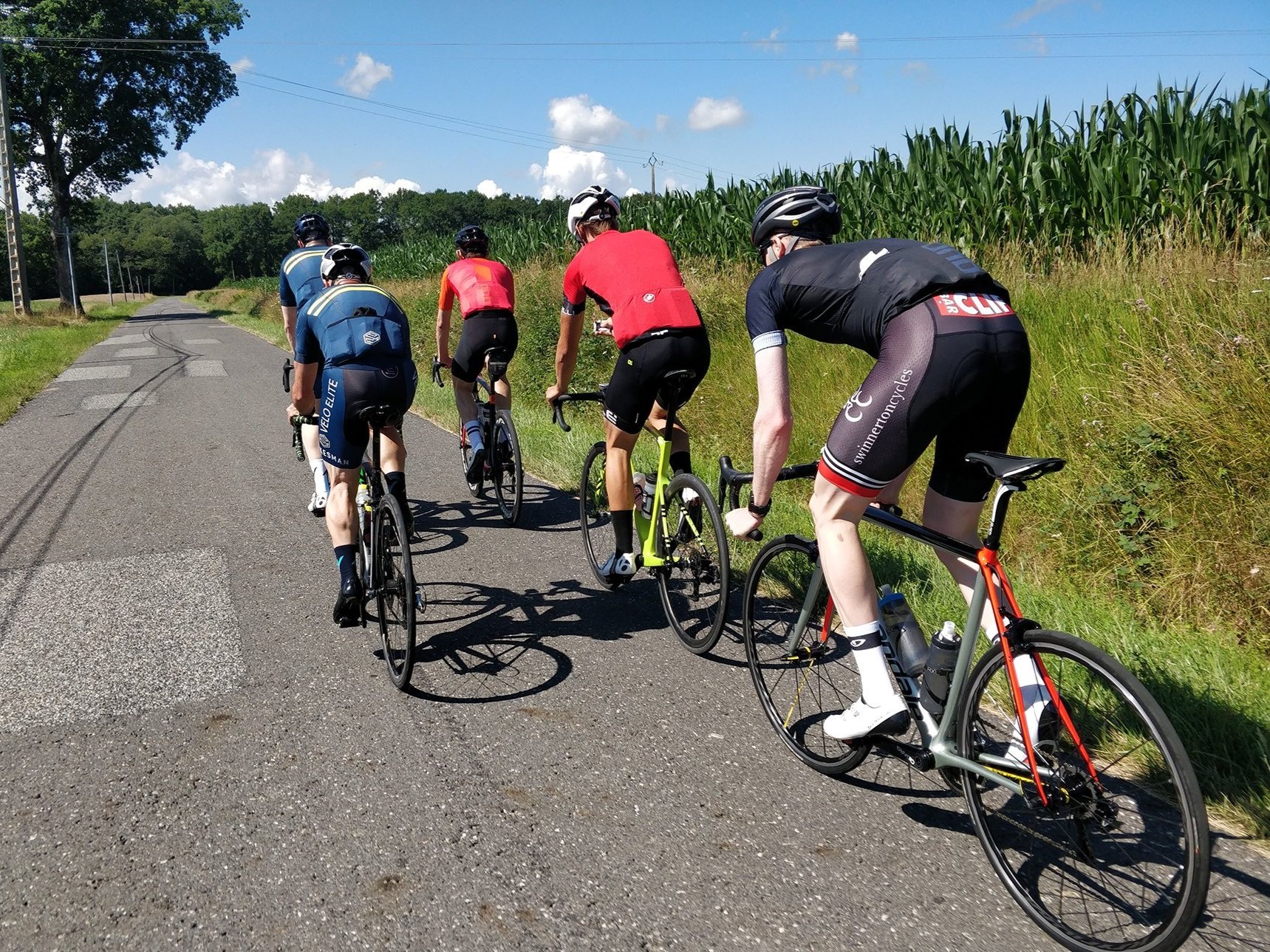 Five cyclists riding their bikes along a rural road surrounded by green fields and blue skies.