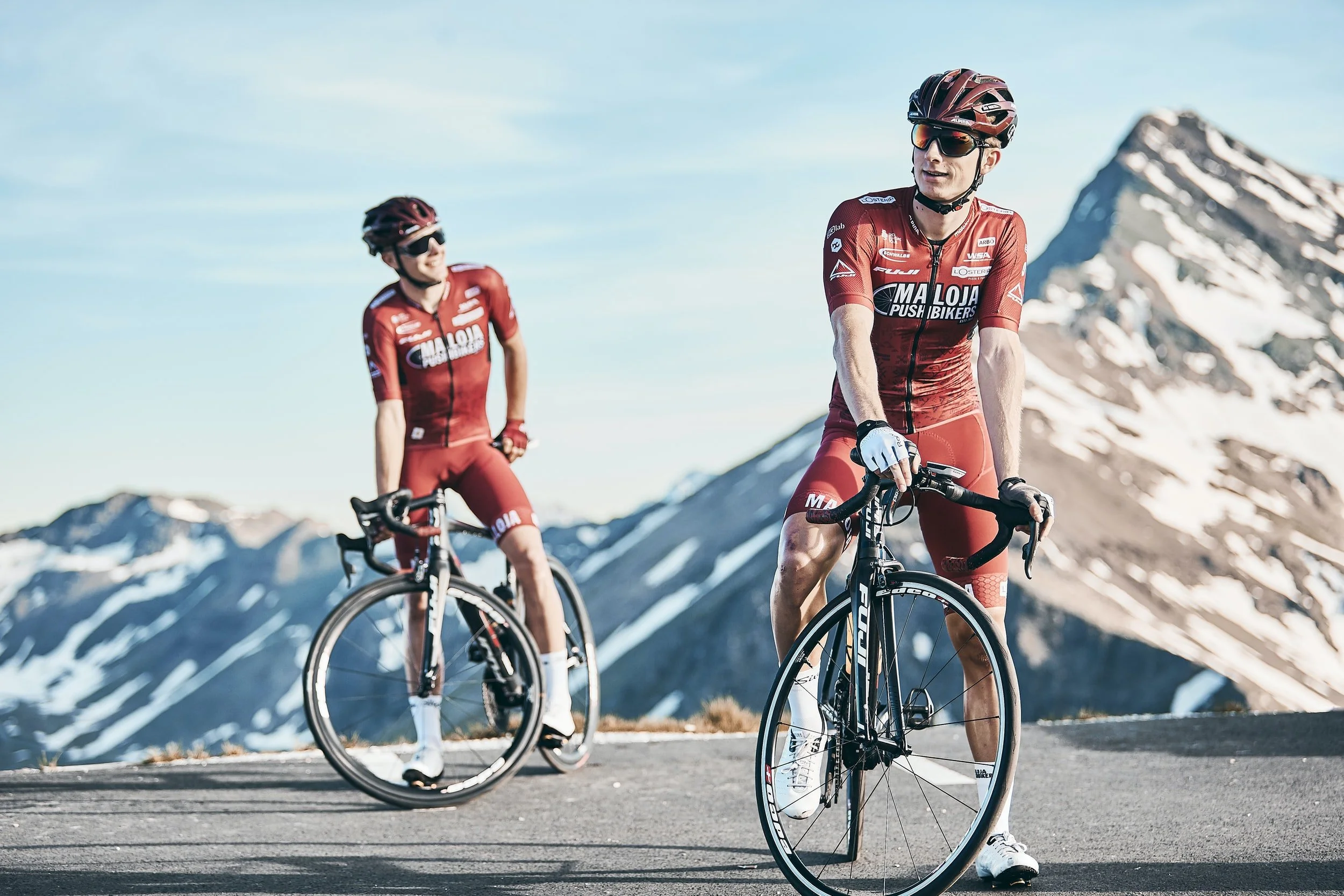 Two cyclists wearing red jerseys and helmets riding bikes on a mountain road with snow-capped peaks in the background.