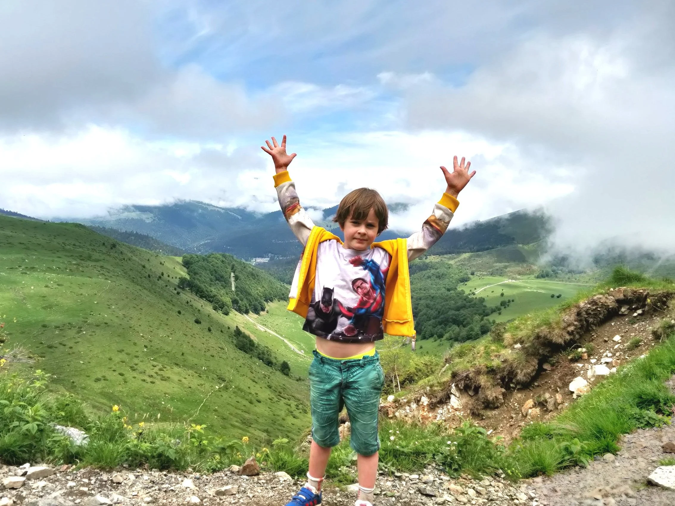 A young boy standing on a hillside with his arms raised in the air, wearing a Batman shirt, shorts, and a yellow jacket, with a green hilly landscape and cloudy sky in the background.