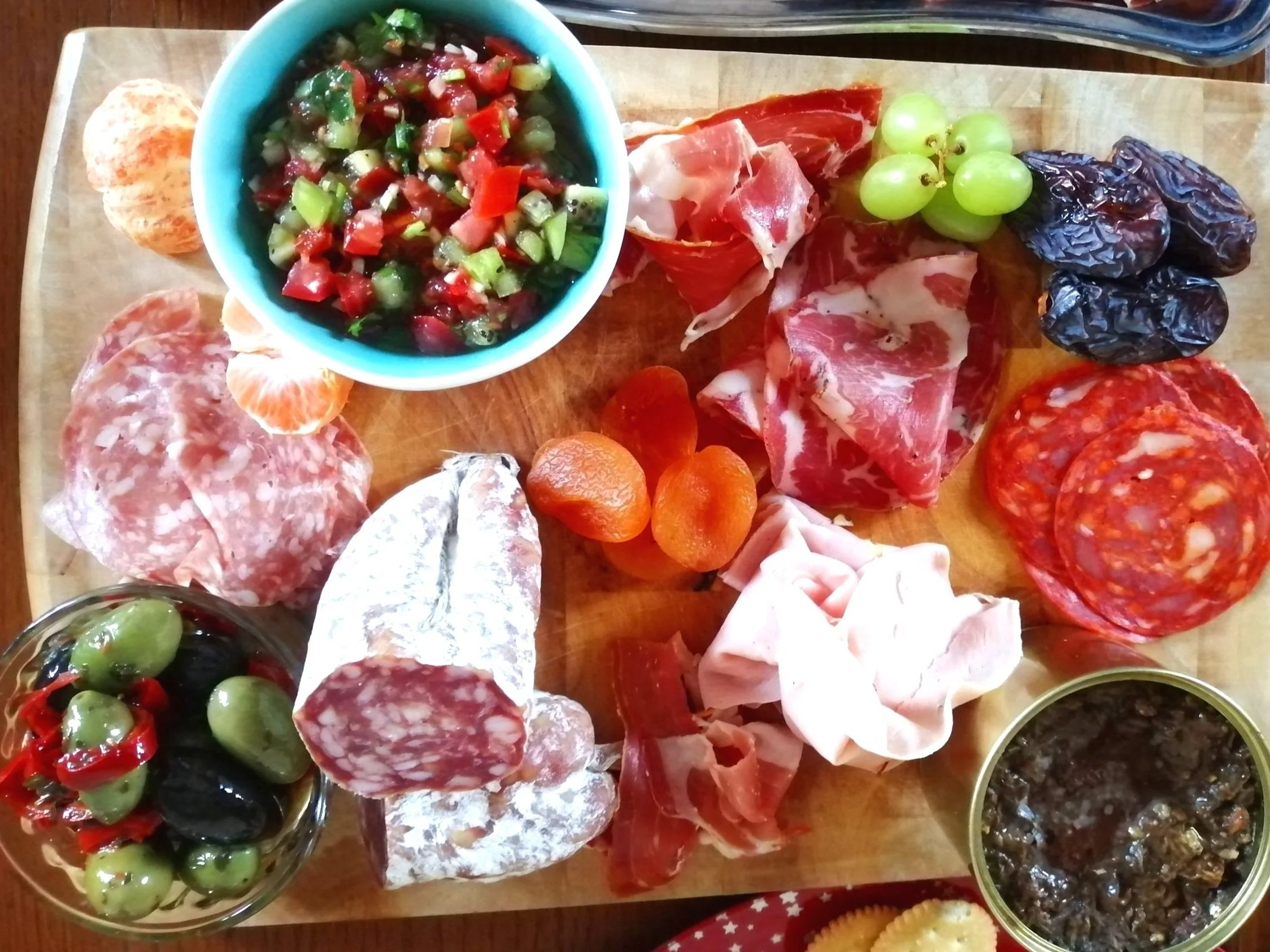 Assorted cured meats, olives, grapes, and a bowl of tomato-based salsa on a wooden serving board.