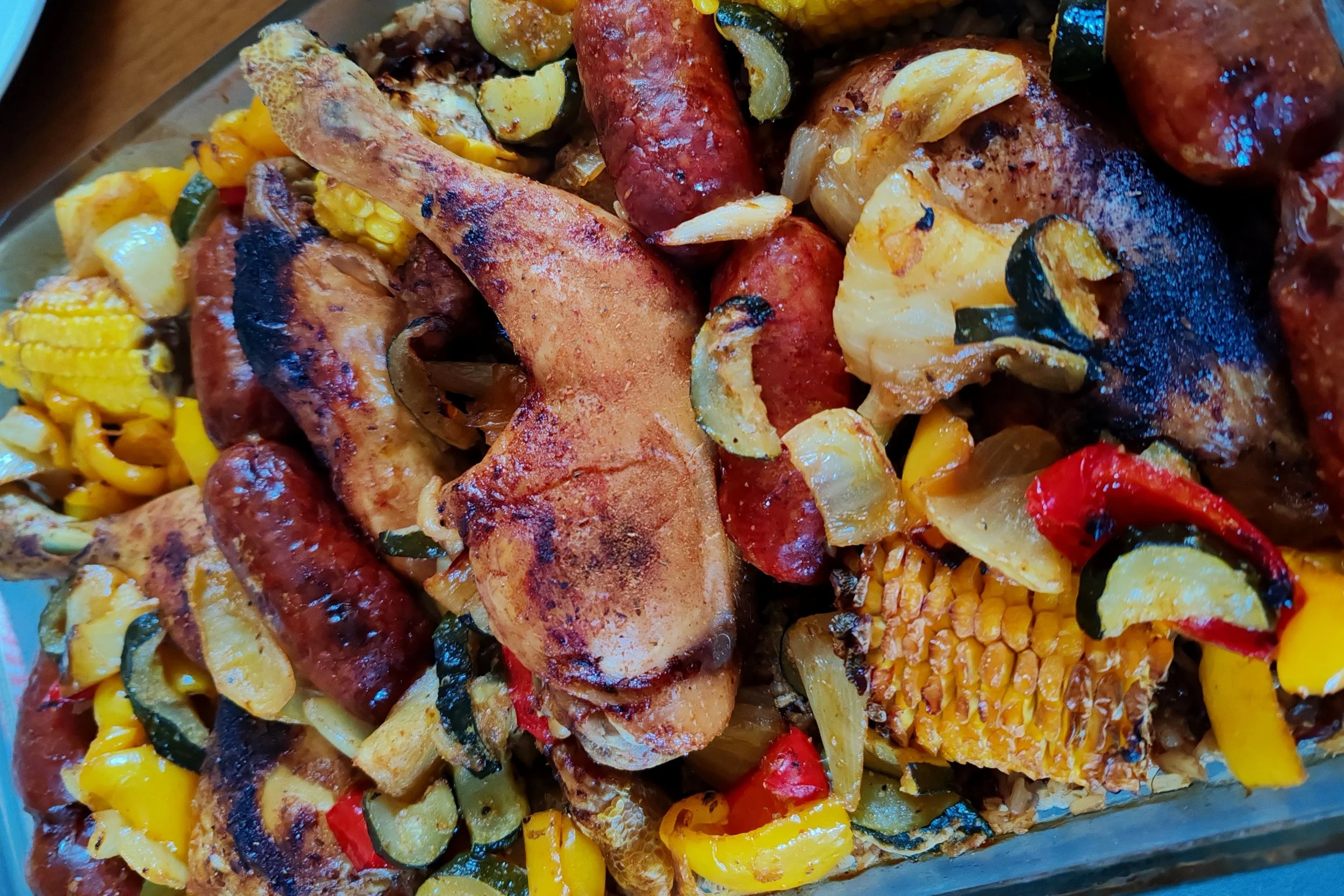 Grilled chicken, sausages, corn on the cob, and roasted vegetables including zucchini, onions, and red and yellow bell peppers.