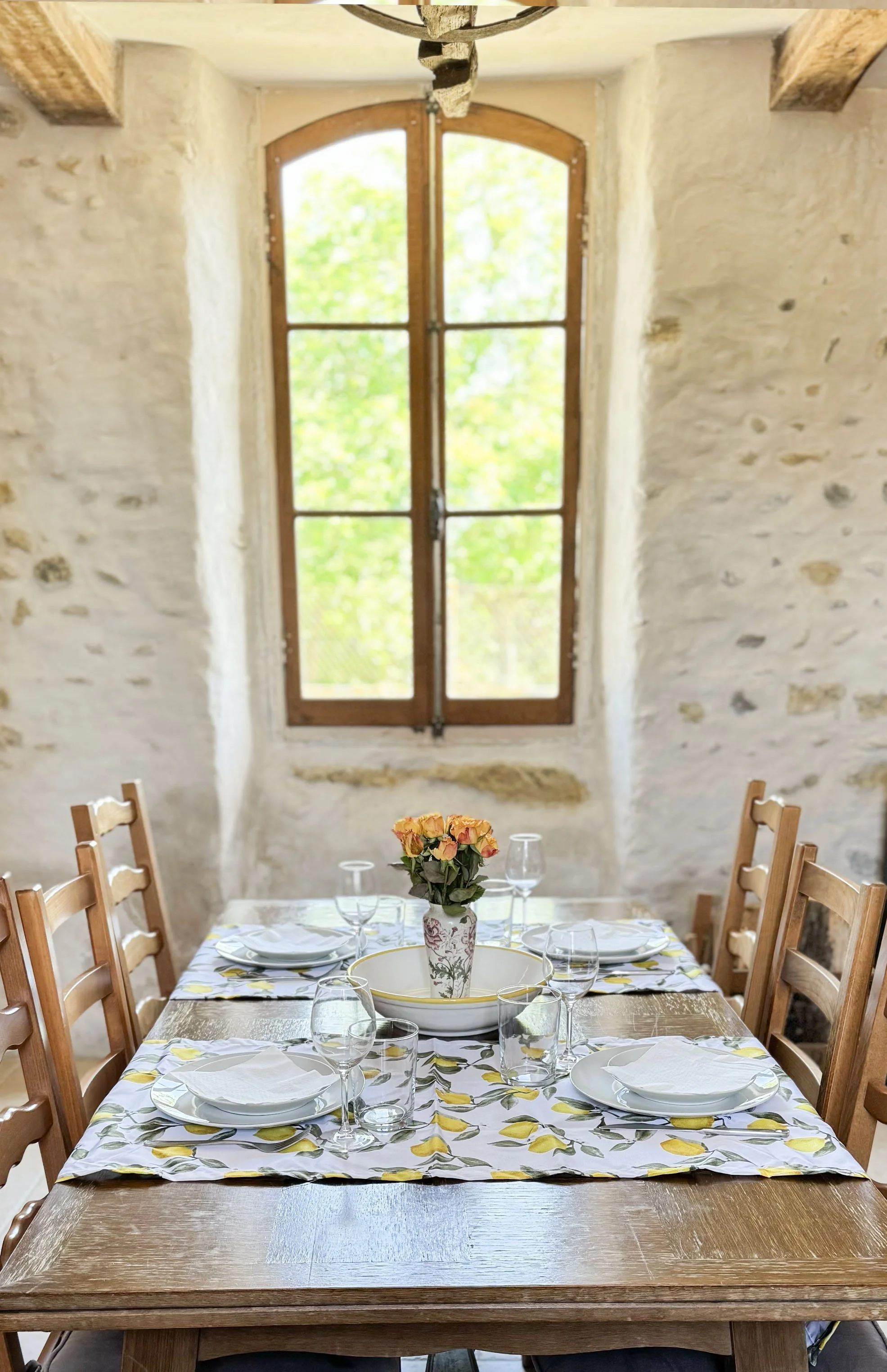 A dining table set for a meal with a floral centerpiece, plates, glasses, and napkins in a rustic room with stone walls and a large window showing greenery outside.