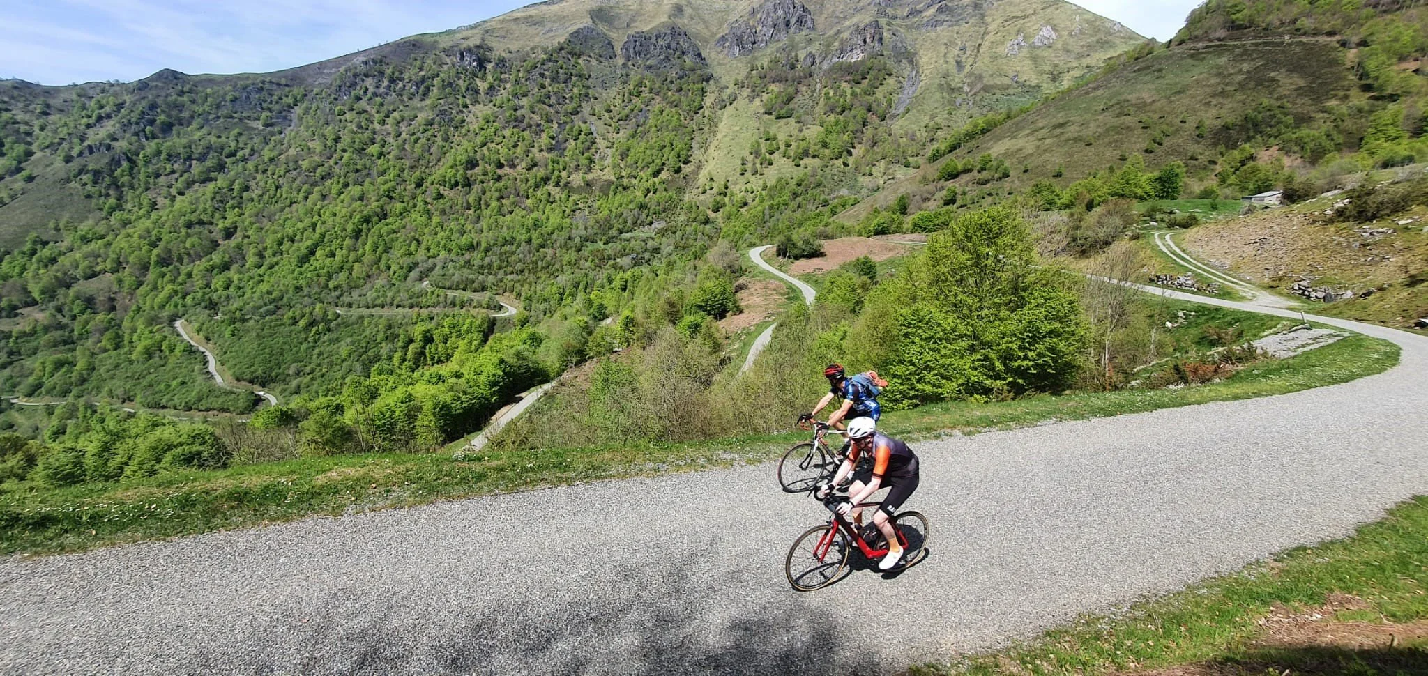Two cyclists riding on a winding mountain road surrounded by green hills and trees.