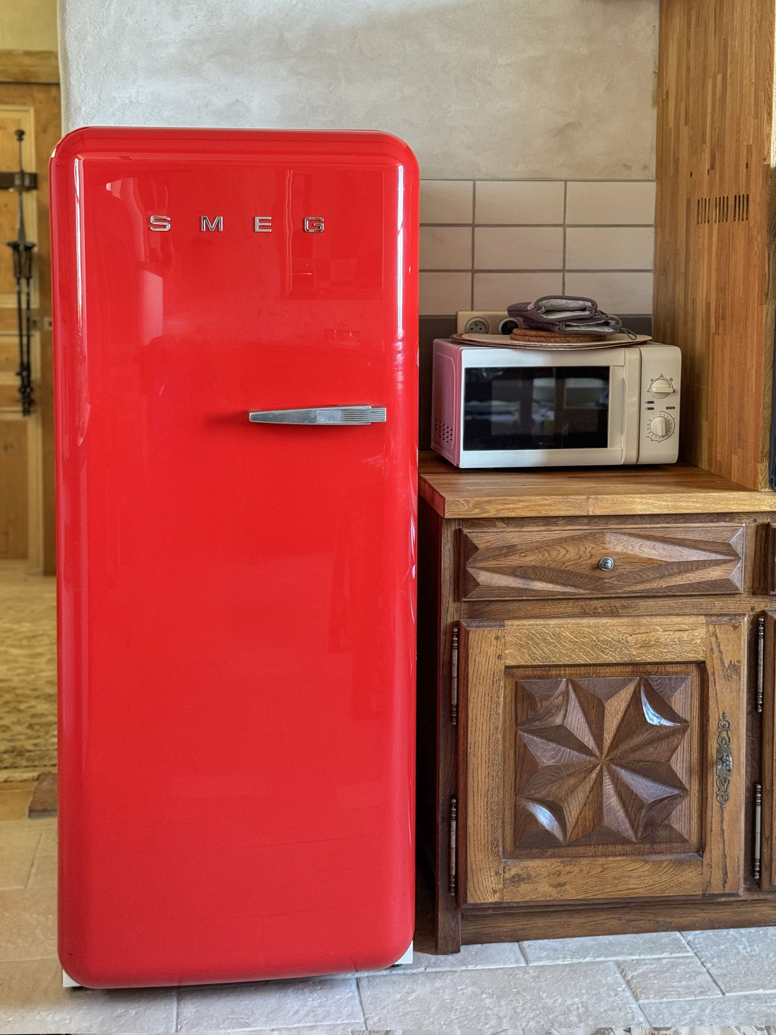 A red Smeg refrigerator in a kitchen next to a wooden cabinet and a microwave oven.