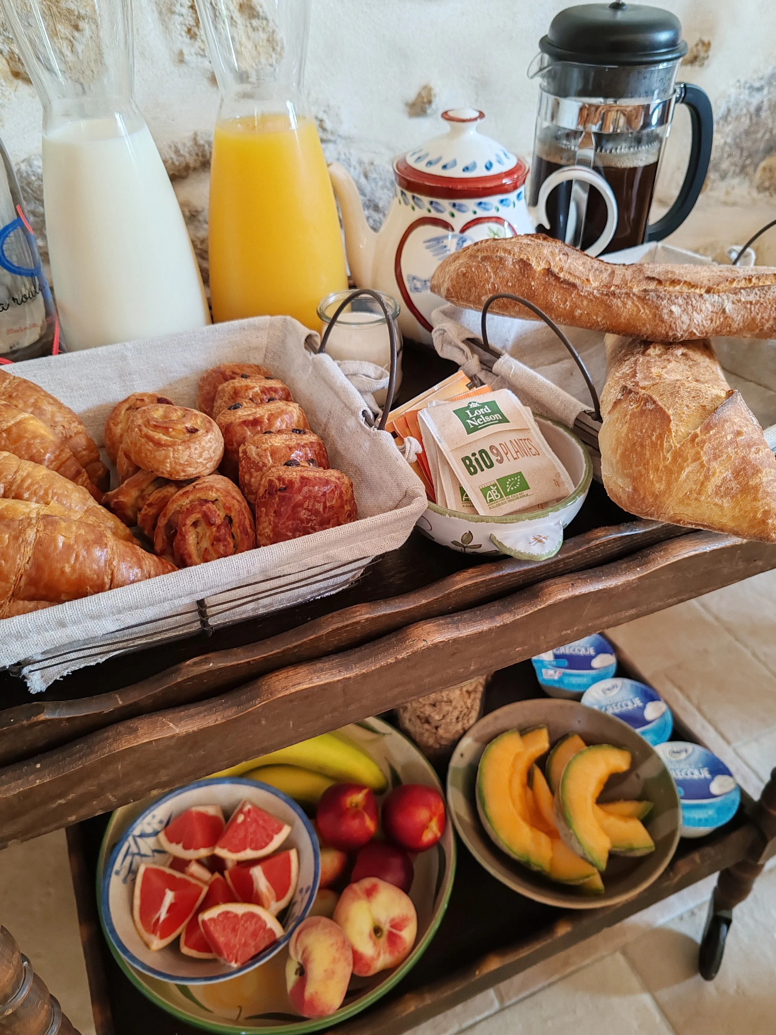 A breakfast display with croissants, sausage rolls, various fruits, bread, and beverages including milk, orange juice, and coffee, arranged on a wooden table against a stone wall.