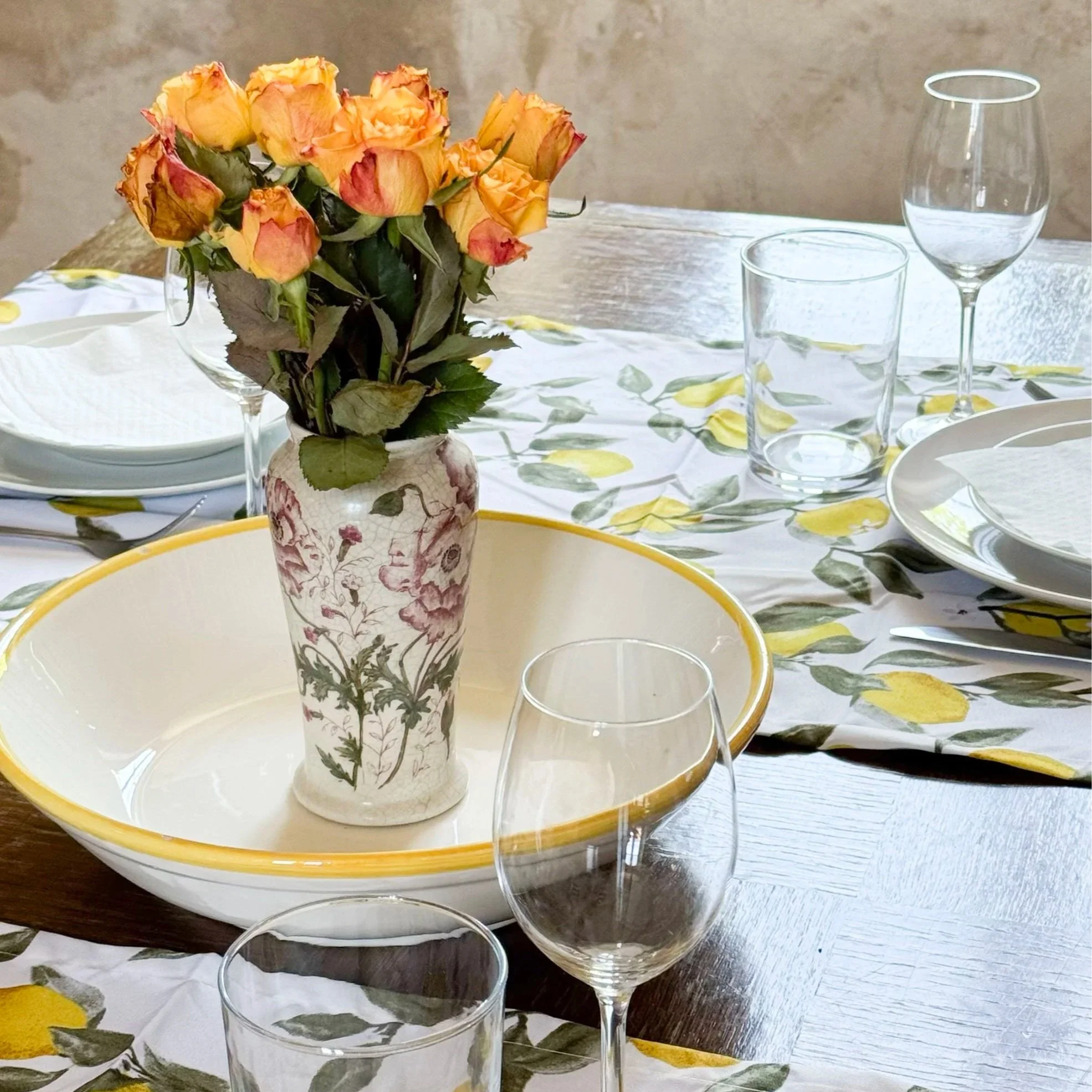 A table set for a meal with a floral theme, featuring a vase of yellow and orange roses, a yellow and white patterned bowl, clear glassware including wine and water glasses, and a white plate with a yellow rim. The tablecloth has a lemon and leaf pattern, and there is a rustic wall in the background.