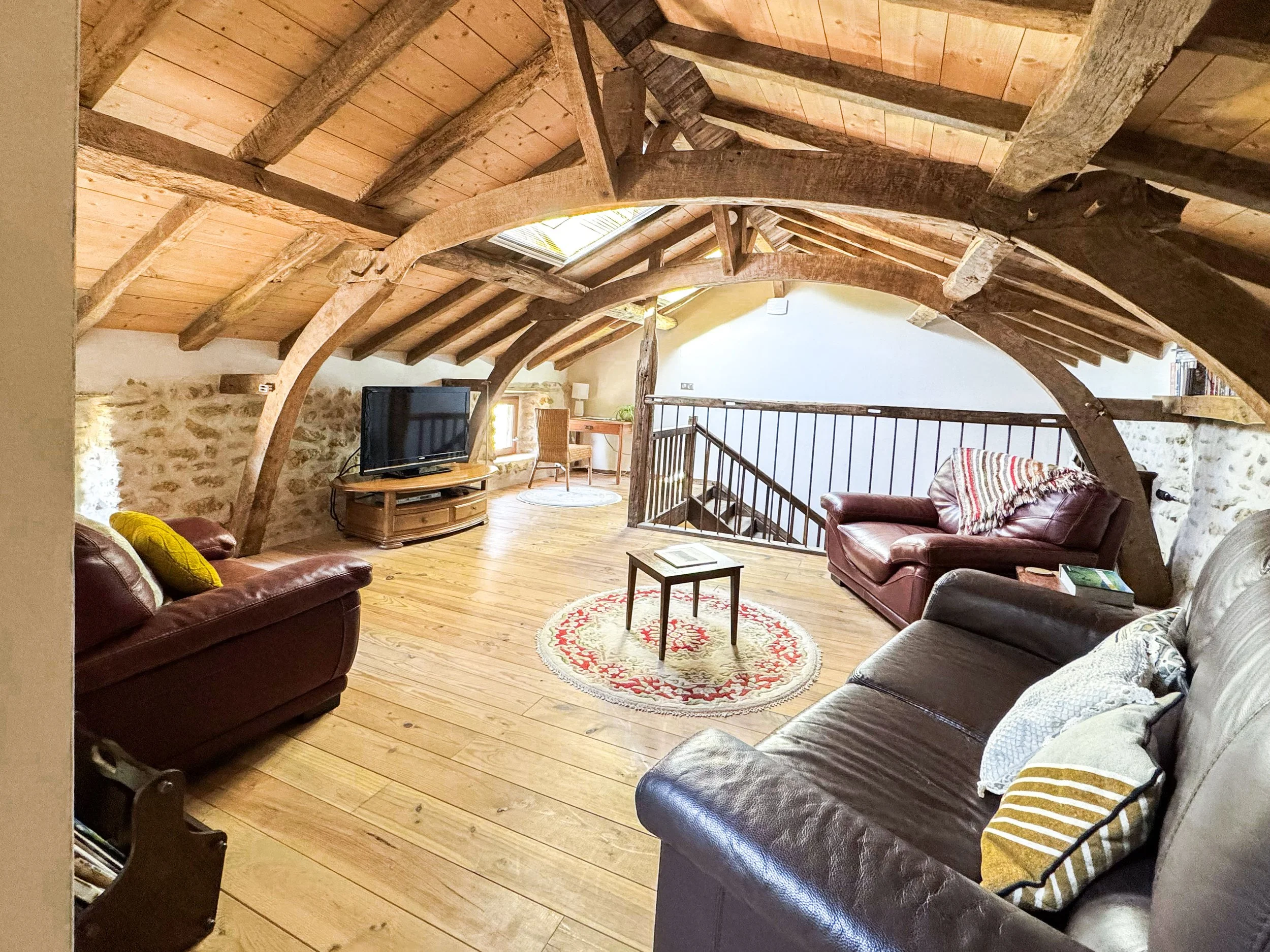 Living room with exposed wooden beams on vaulted ceiling, stone wall, hardwood floors, leather sofas with striped and yellow pillows, a round rug, a wooden coffee table, a flat-screen TV, a wicker chair, and railing indicating a loft or balcony area.
