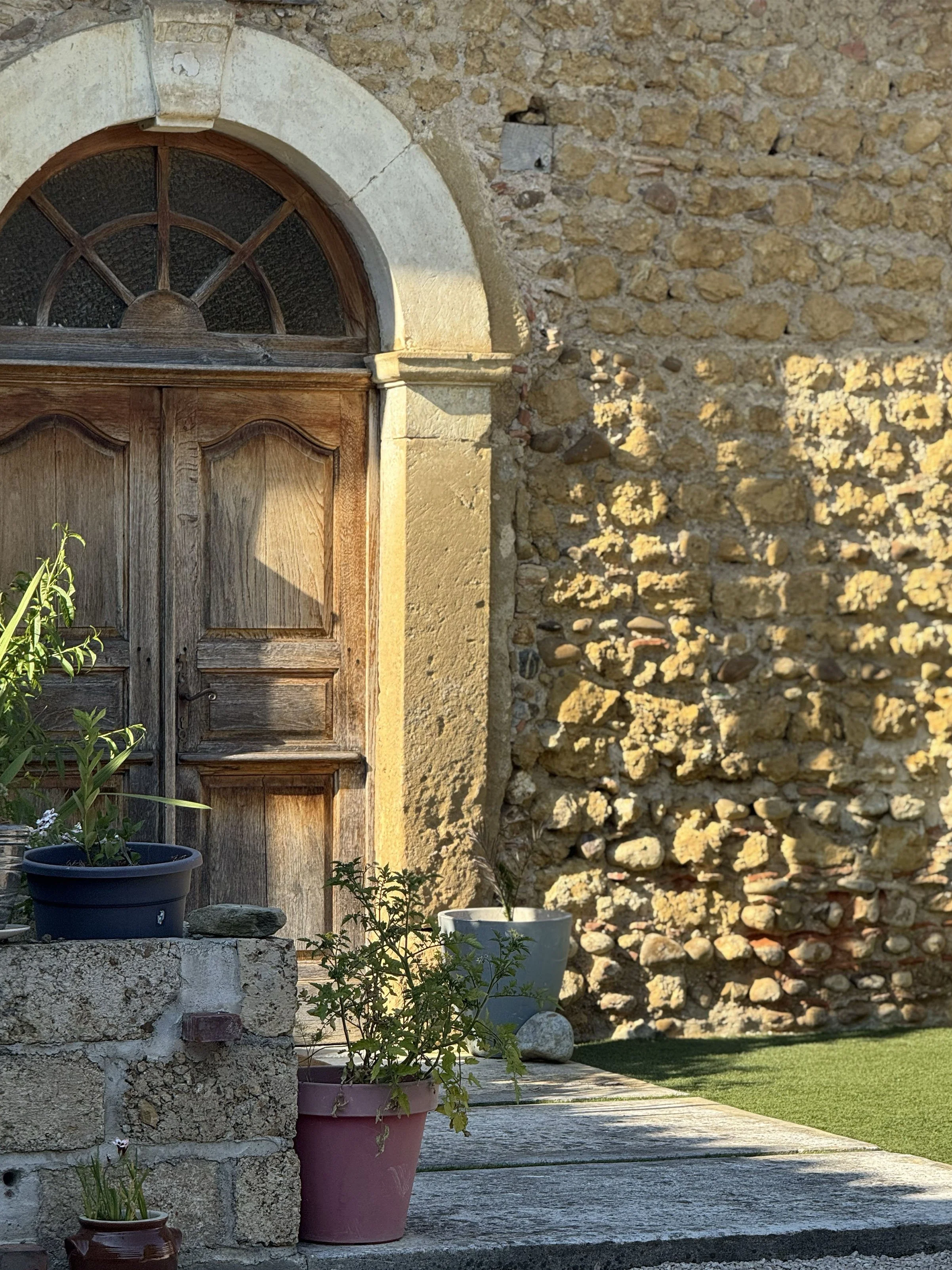 A stone wall with a wooden arched window and potted plants on a stone step leading up to it, in sunlight.