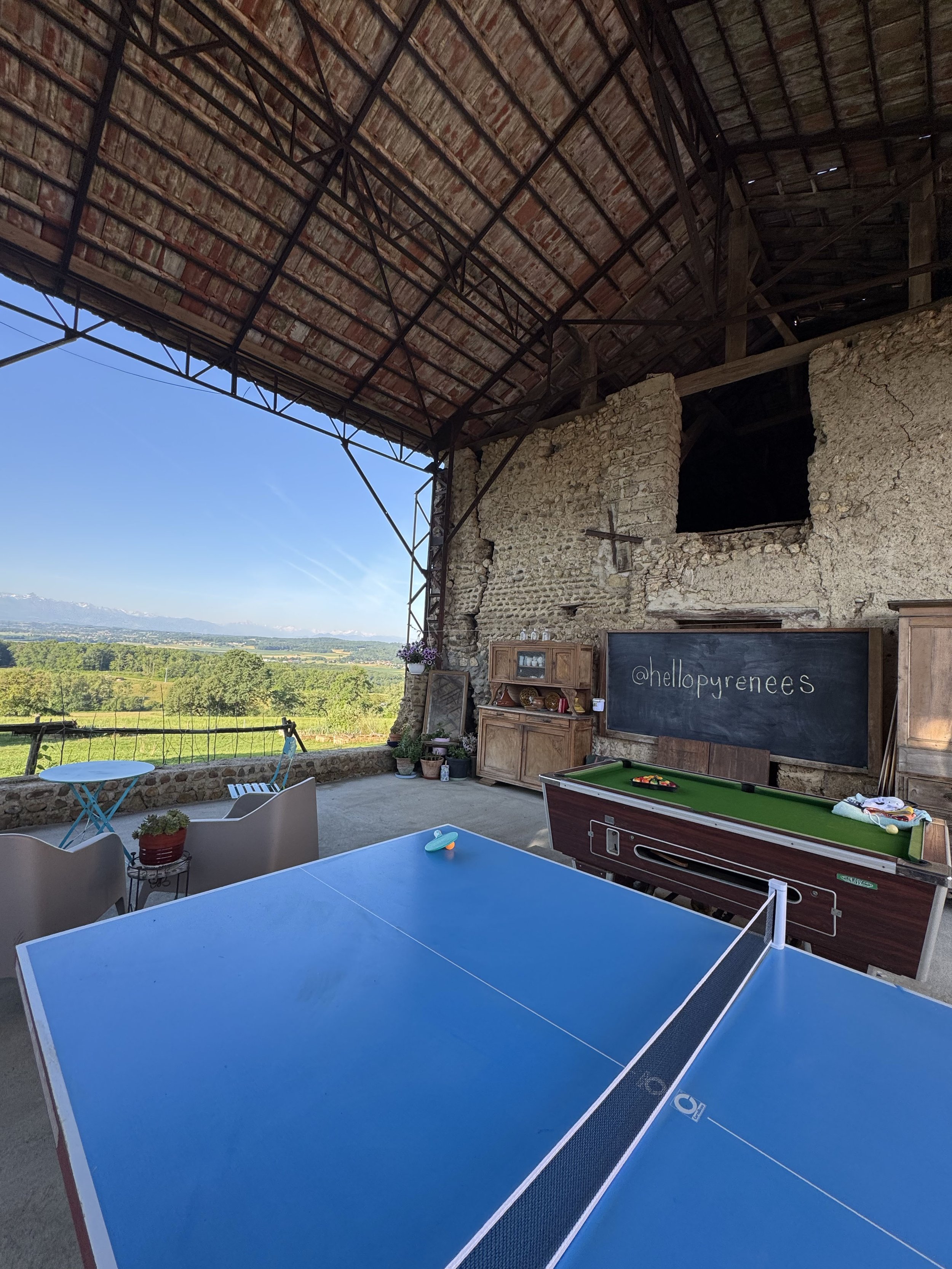 View from a covered patio showing a blue ping pong table, a pool table, and an outdoor scenic landscape with green fields and mountains in the distance.