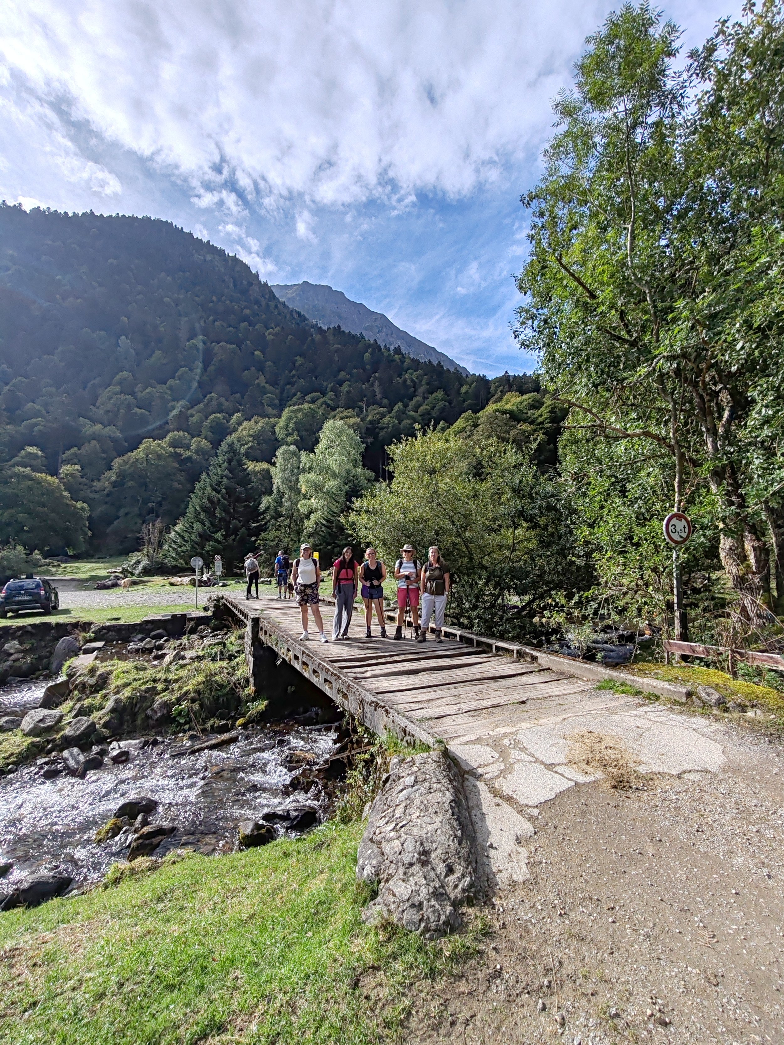 Group of hikers crossing a small wooden bridge over a stream in a wooded mountain landscape with a partly cloudy sky.