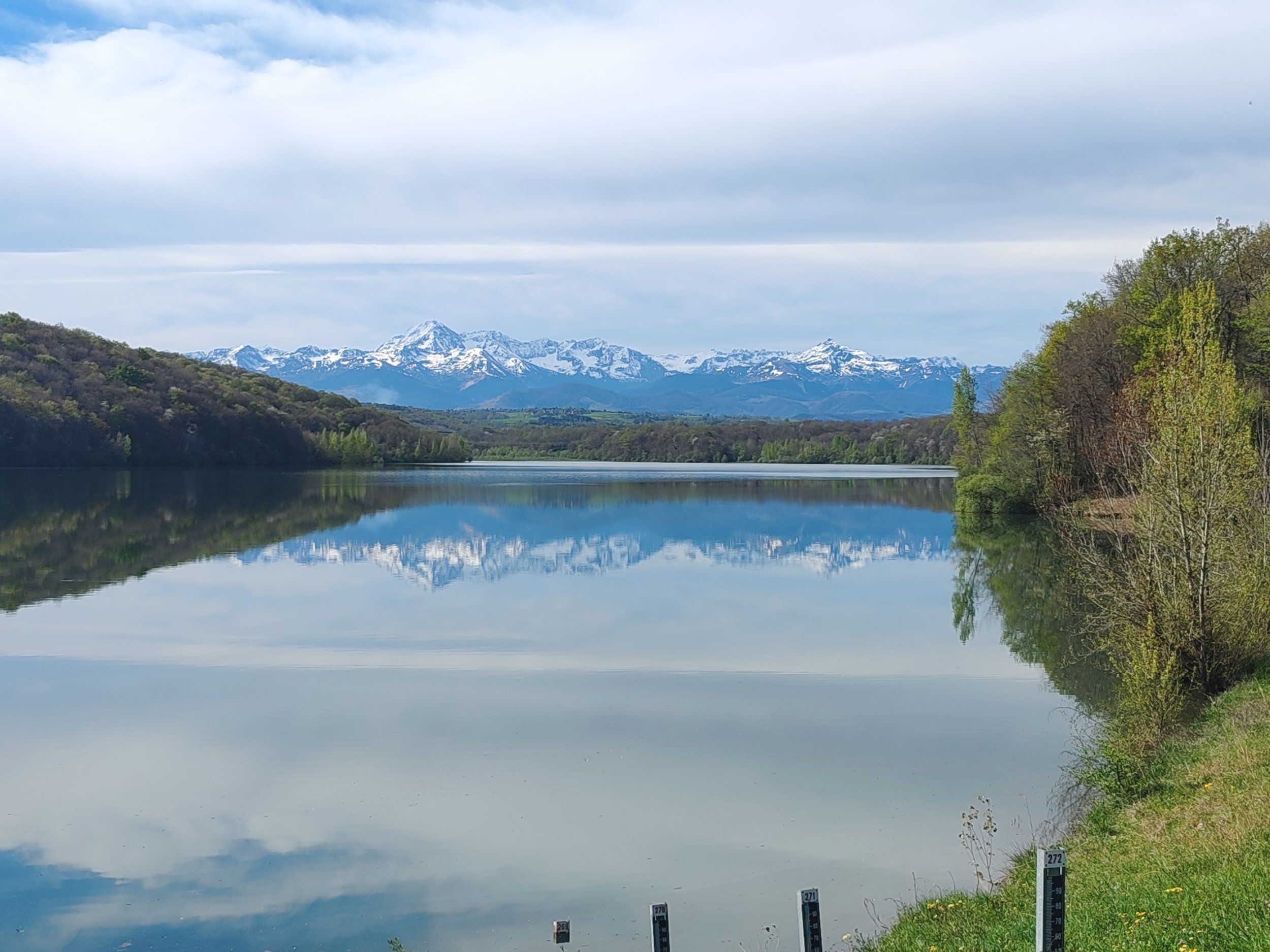 A calm lake reflecting snow-capped mountains and cloudy sky, with green trees lining the shoreline.