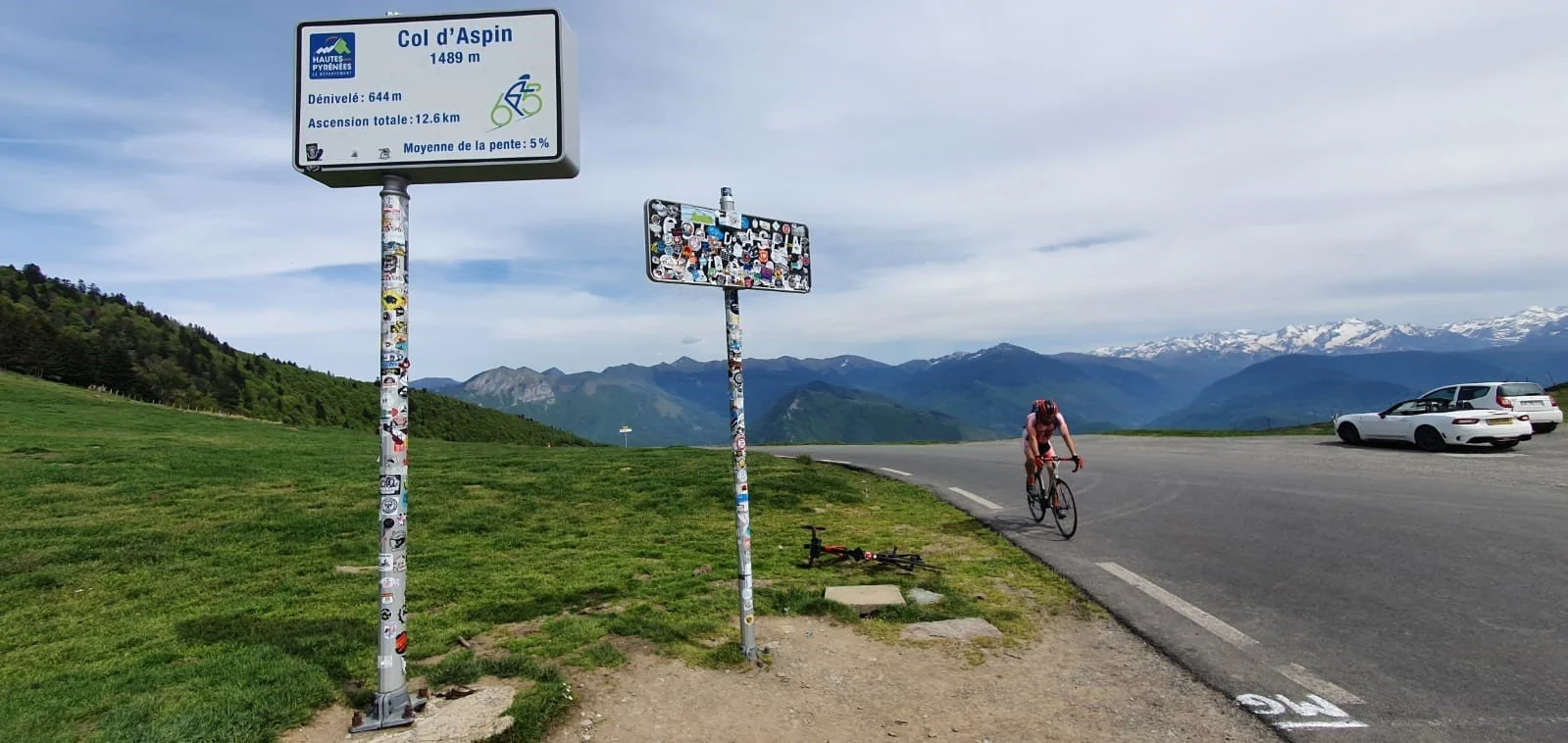 A cyclist riding uphill on a mountain road near the Col d'Aspin at 1489 meters elevation, with green hills and snow-capped mountains in the background. There are parked cars and signs next to the road, one of which indicates the elevation and other cycling info.