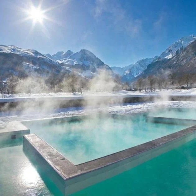 Hot spring with steaming water outdoors, surrounded by snow-covered mountains under a clear blue sky with the sun shining.