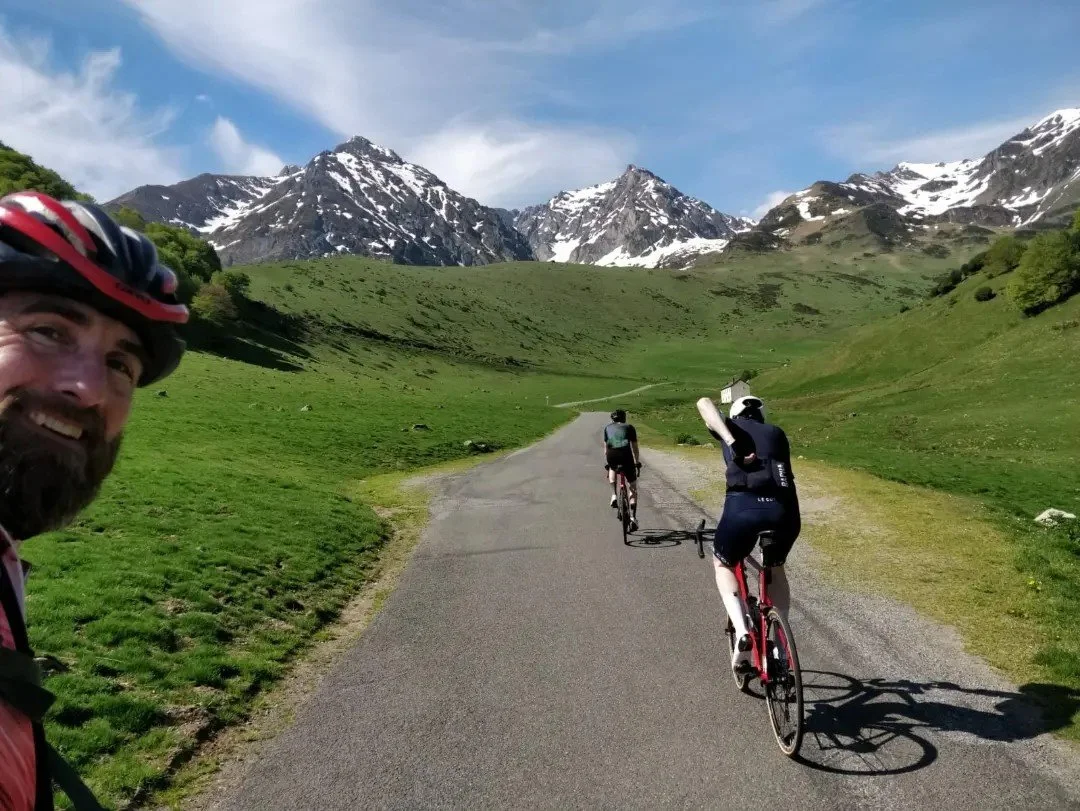Three cyclists riding on a paved trail through a green mountainous landscape with snow-capped peaks under a partly cloudy sky.