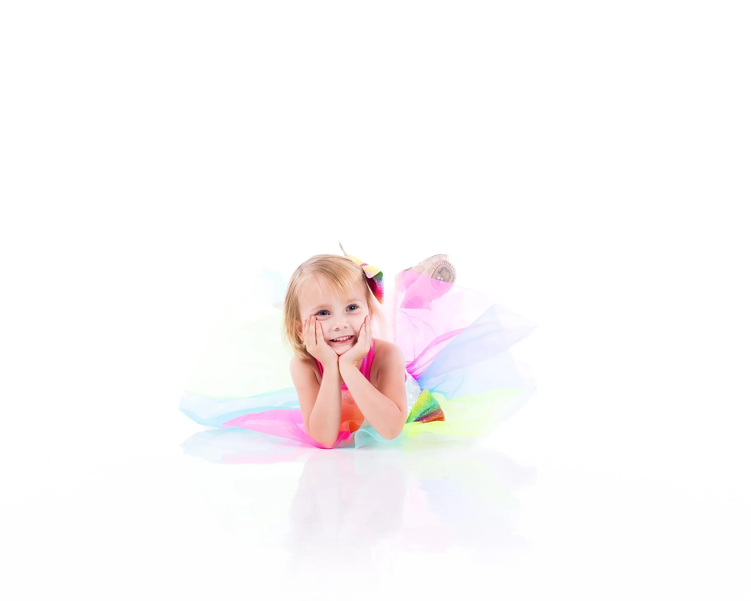 A young girl lying on her stomach, smiling with her hands on her cheeks. She is dressed in a colorful, frilly dress with rainbow-colored accents, and she has a rainbow-colored hair bow.