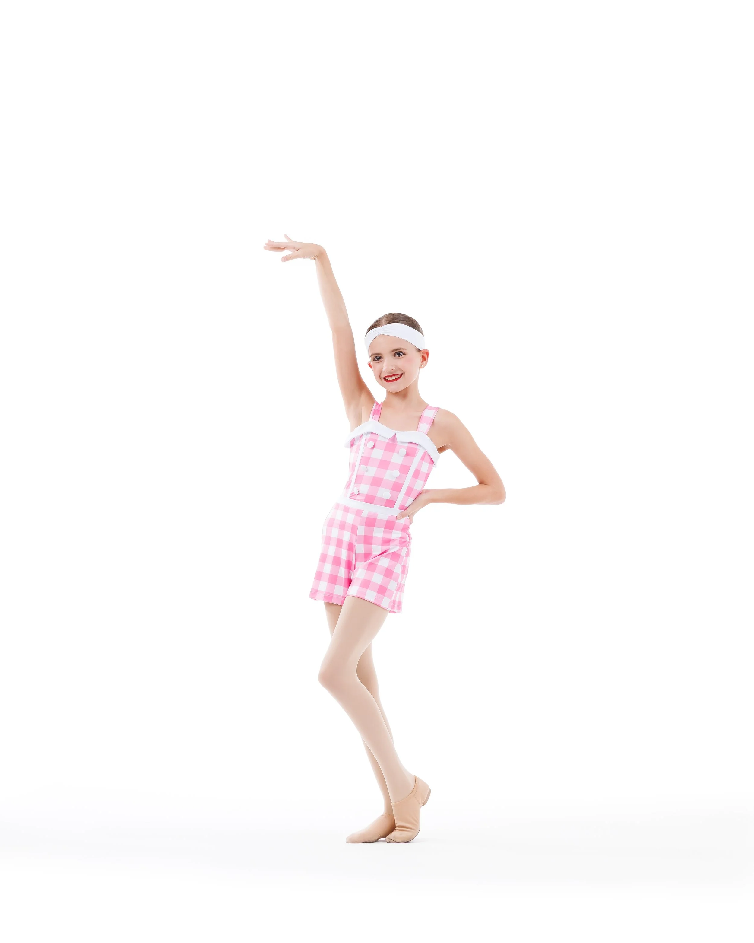 A ballerina in pink checkered dance attire, white headband, and pointe shoes, standing on one leg with the other slightly raised, smiling with her right arm raised.