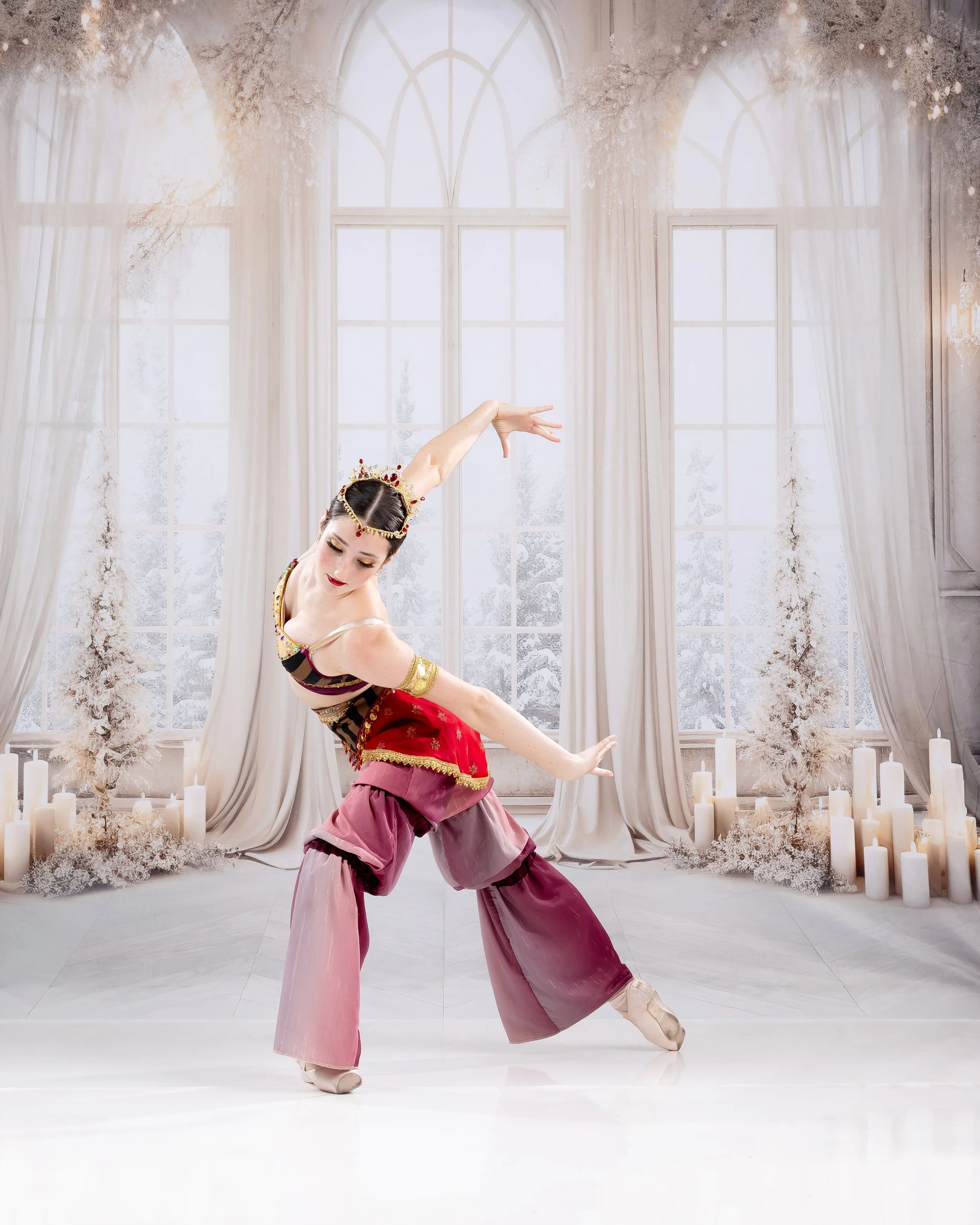 A female ballet dancer in a colorful costume performing a dance pose in a bright room with large windows and white candles.