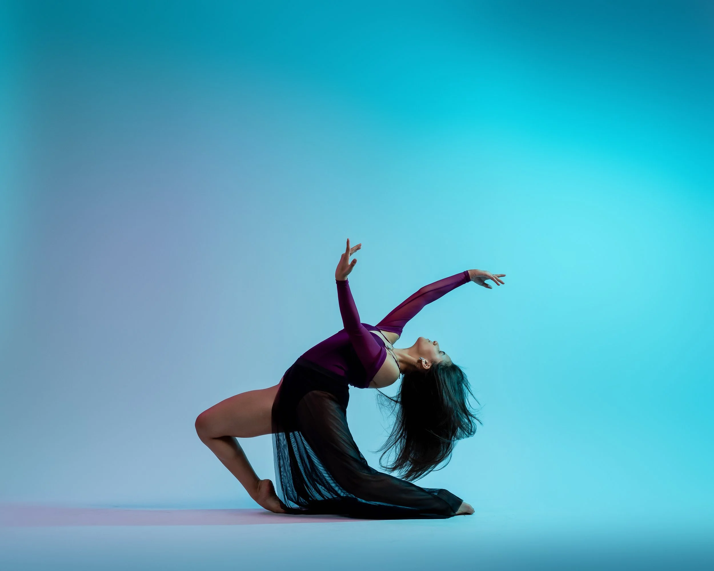 A woman in a purple top and black sheer skirt performs a dance pose against a light blue background, with her body curved and her arms extended.