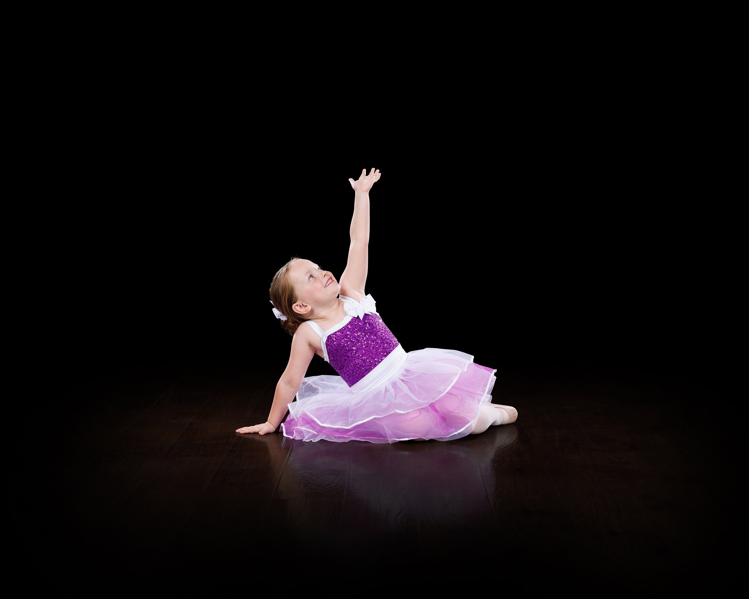 Young girl in a purple ballet dress sitting on the floor and reaching upward with her right arm against a black background.