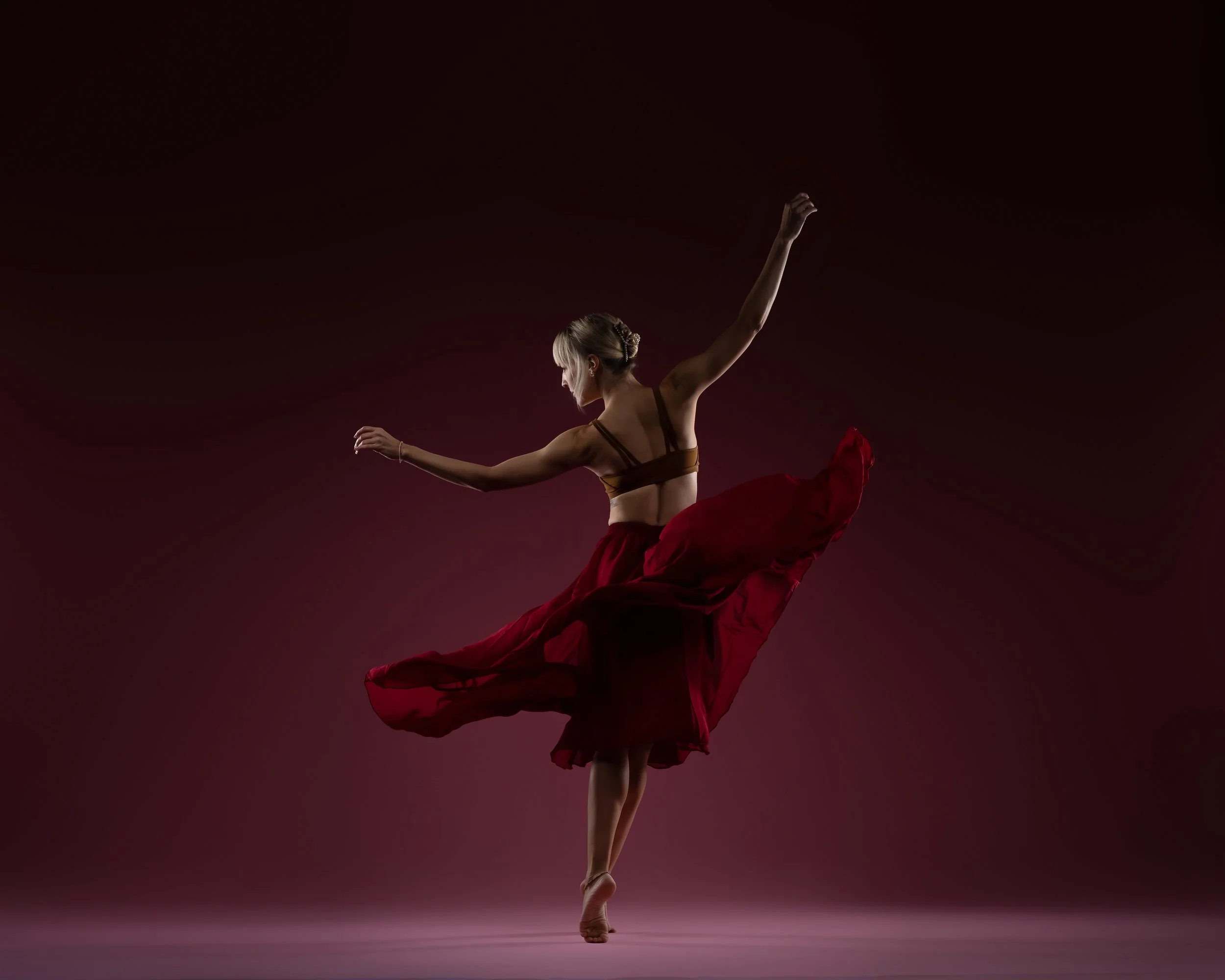 A ballet dancer in a pose with arms extended and one leg lifted, wearing a red skirt and a brown top, against a dark background.