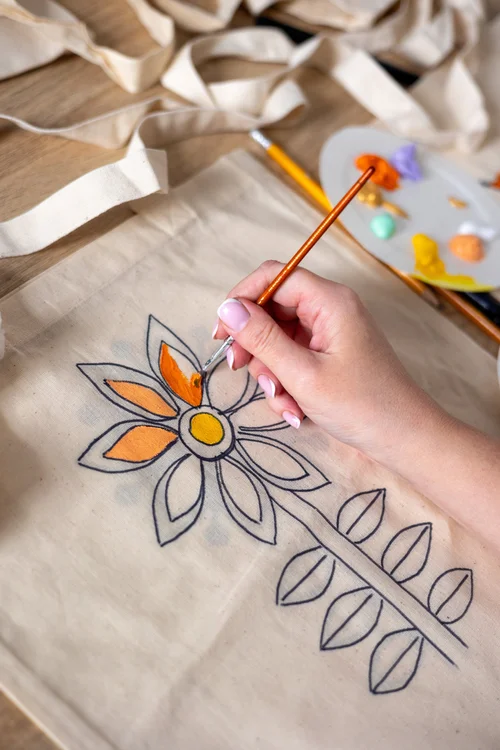 Hand painting an orange and yellow flower design on a canvas tote bag with a paintbrush and palette nearby
