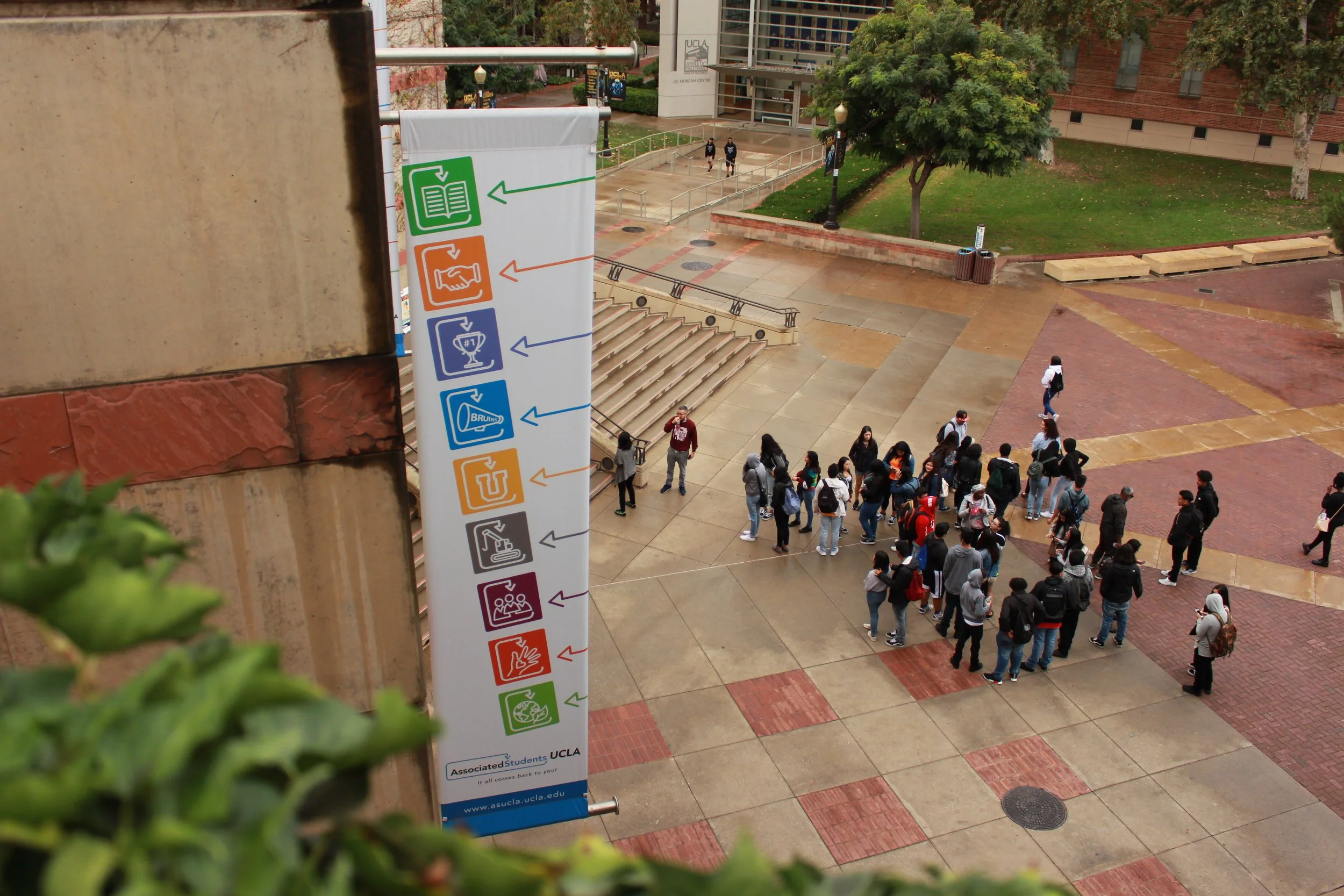 White banner with colorful symbols attached to side of building hangs above people on pathway below.