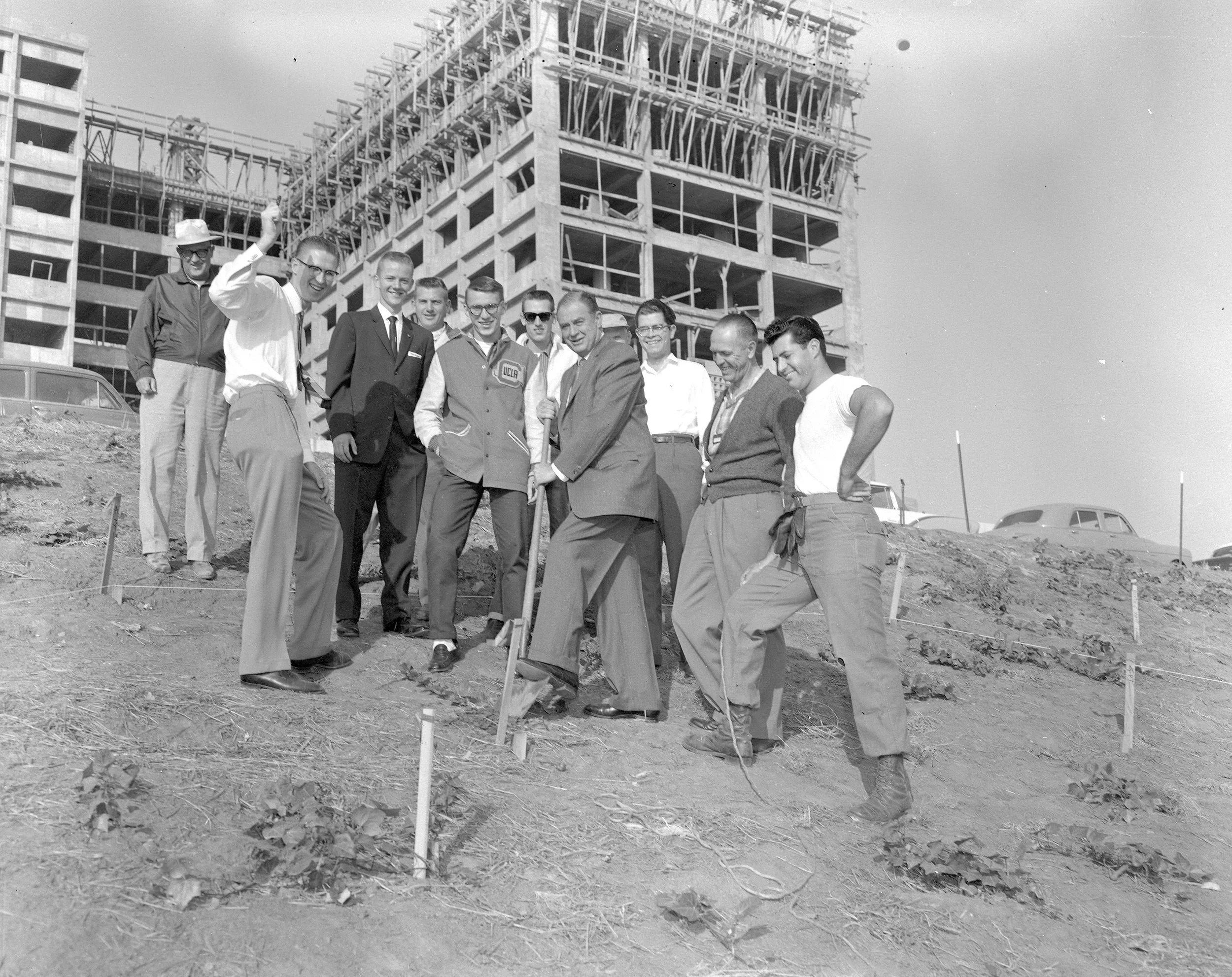 Group of men stand on dirt hill in front of a half-constructed building. One man holds a shovel in the ground.