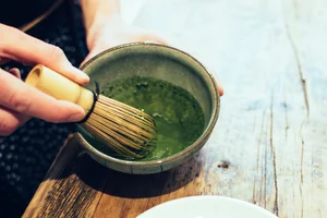 Person using a bamboo whisk to prepare green matcha tea in a ceramic bowl on a wooden table