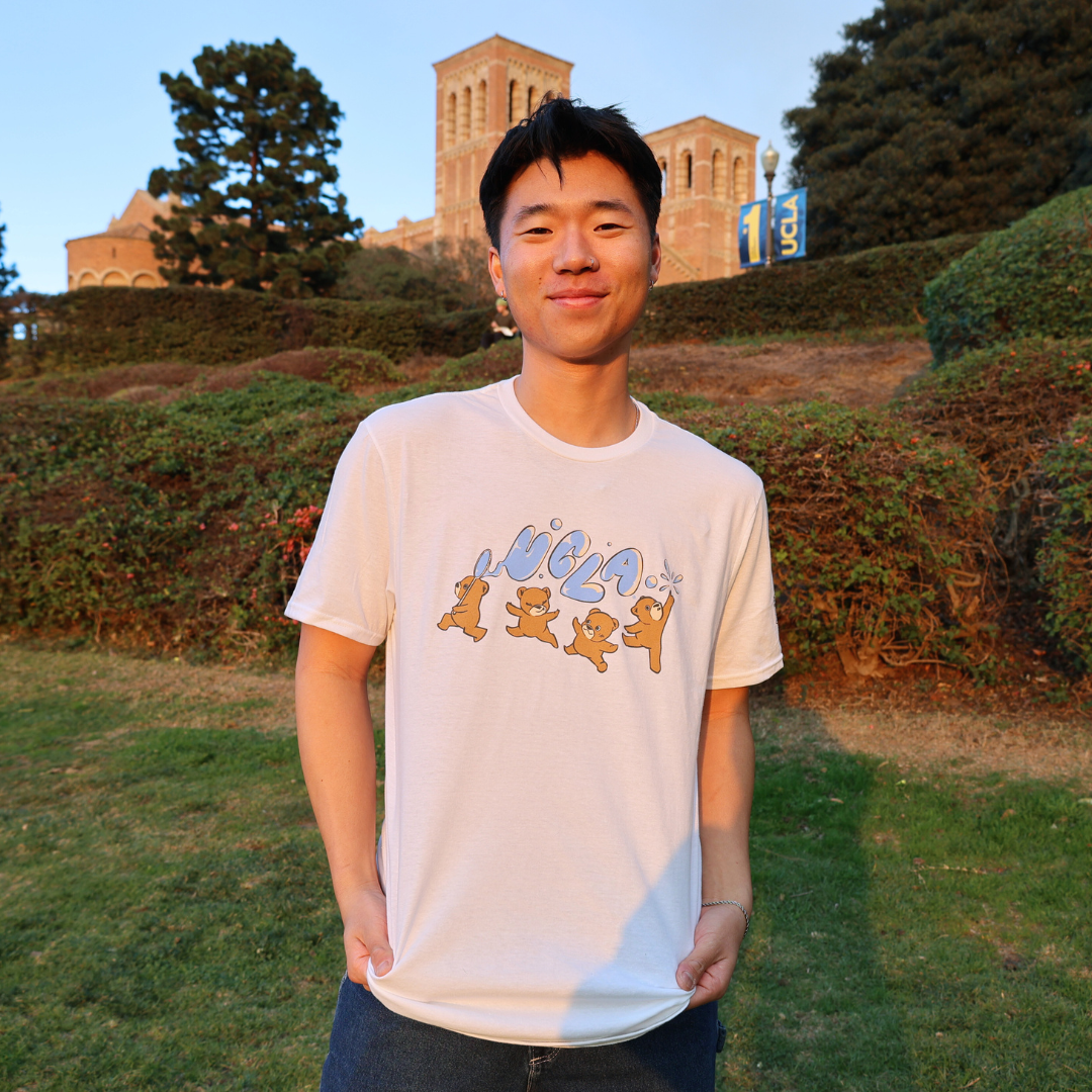 Man wearing the winning T-shirt design featuring bears holding bubbles that spell “UCLA,” standing on a grassy lawn with a campus building in the background.