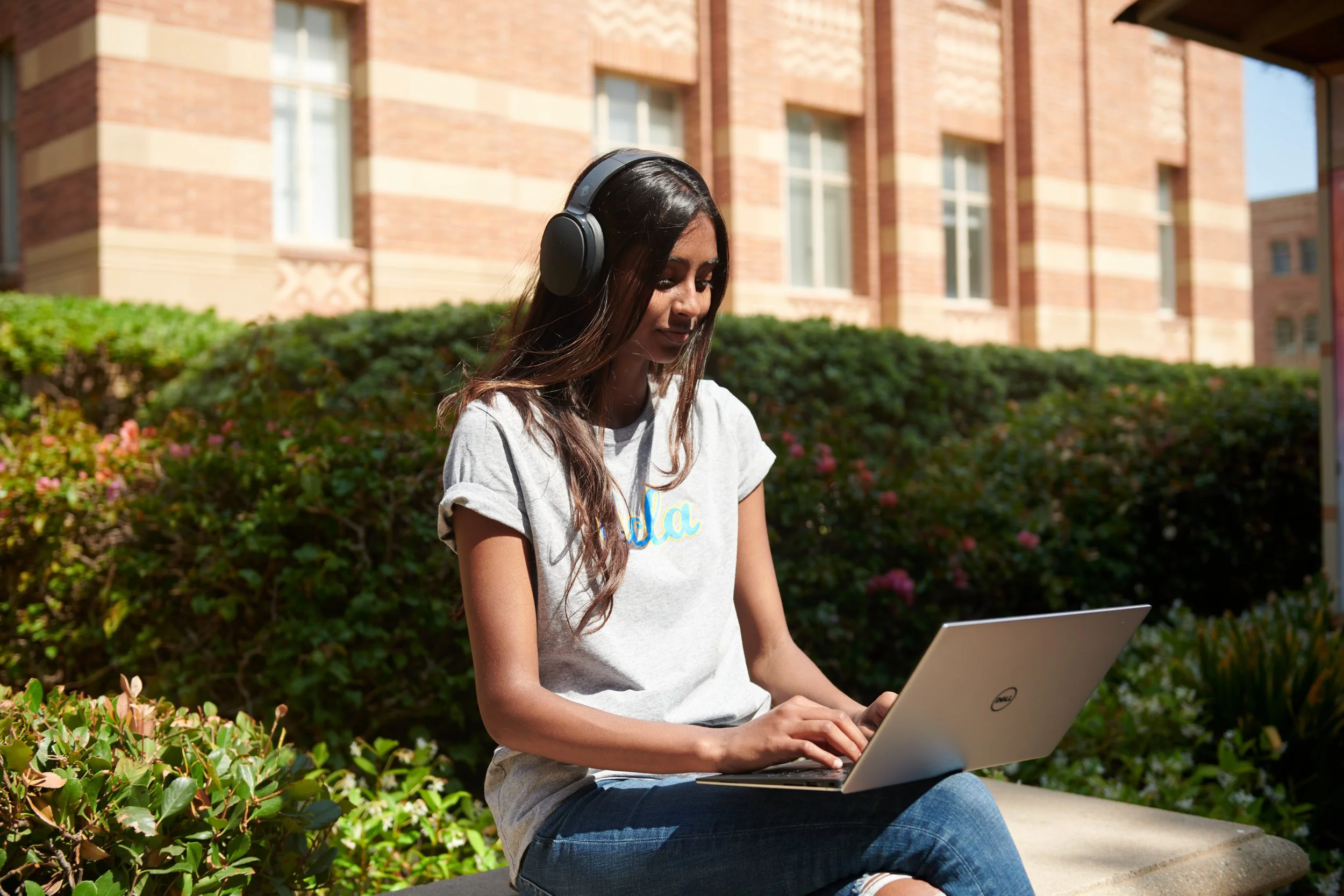 A female student wearing headphones and typing on her laptop sitting on a bench in front of Royce Hall