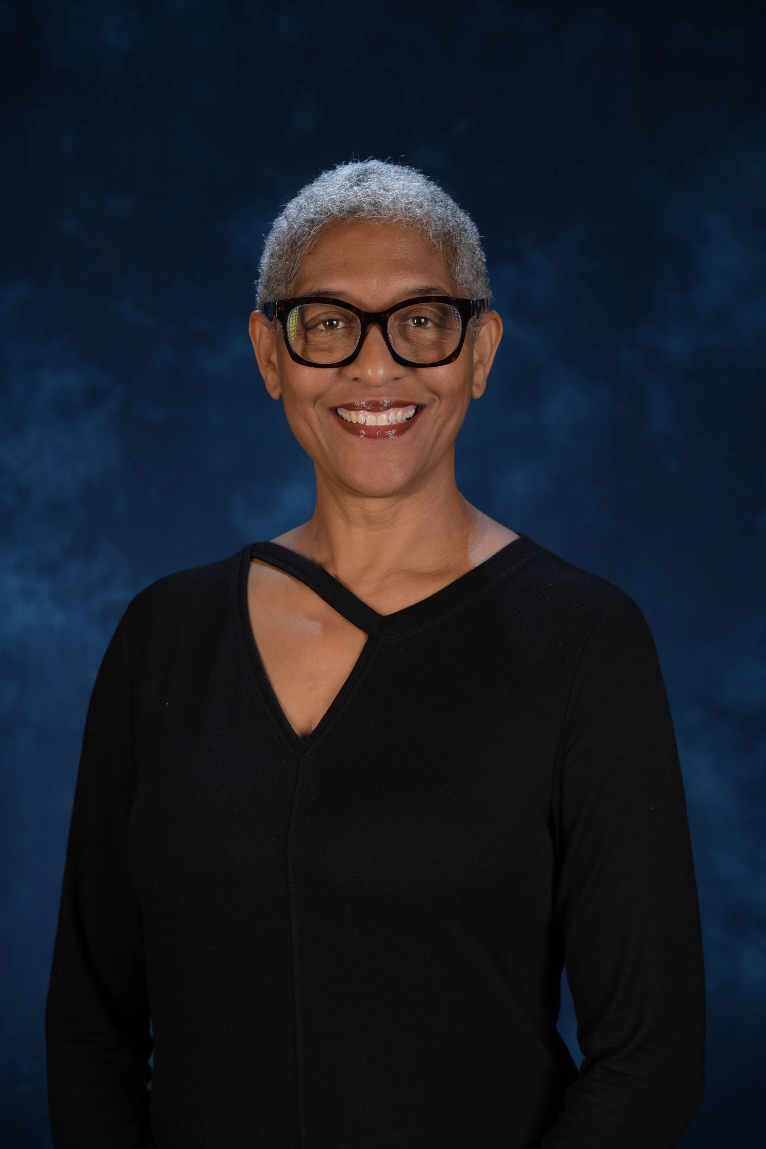 Professional portrait of a smiling woman with short hair and glasses, wearing a black blouse against a dark blue background.