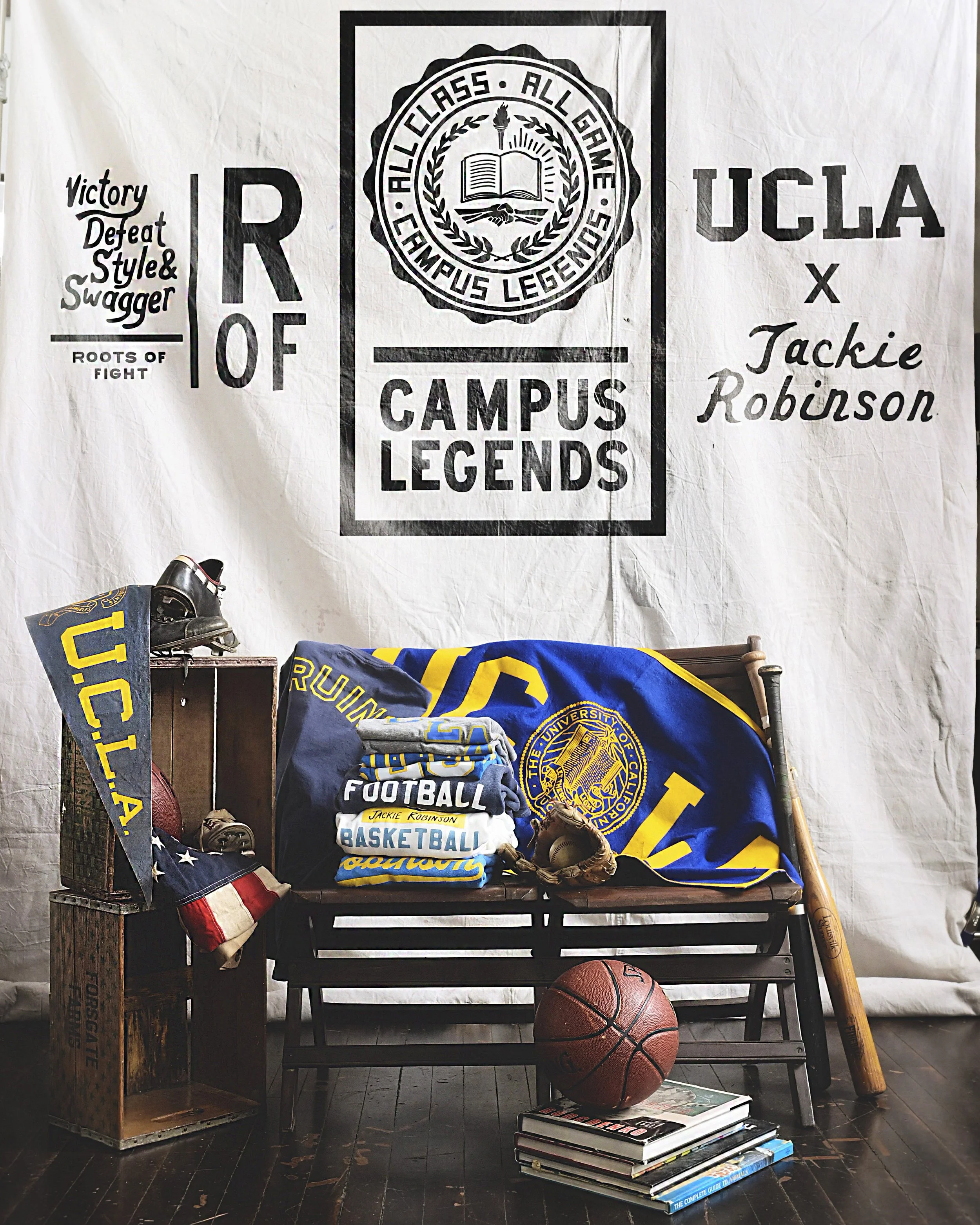A stack of UCLA athletic apparel and merchandise propped on chairs with other athletic accessories in front of a banner for the Campus Legends, UCLA X Jackie Robinson collection.