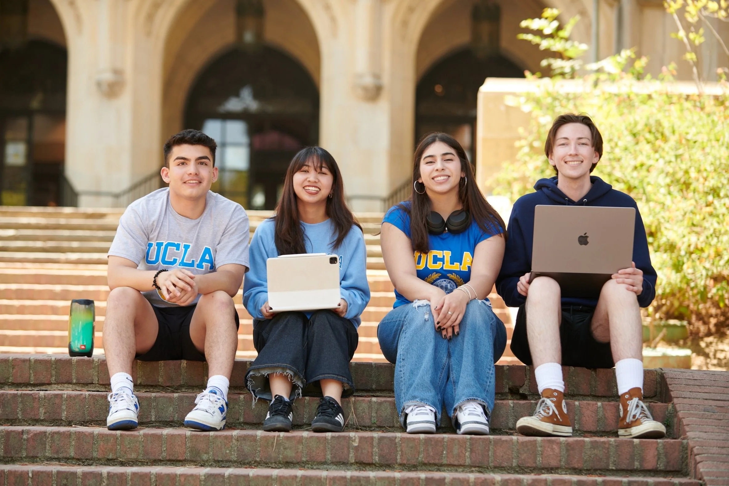 Four students sitting in a row on steps, featuring a speaker, iPad and laptop.