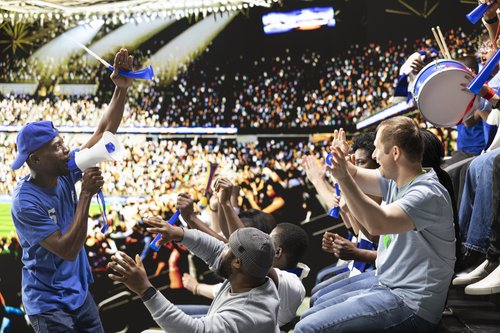 Students cheer together during a lively sports watch party, waving blue noisemakers, clapping, and playing a drum in front of a large stadium-style screen