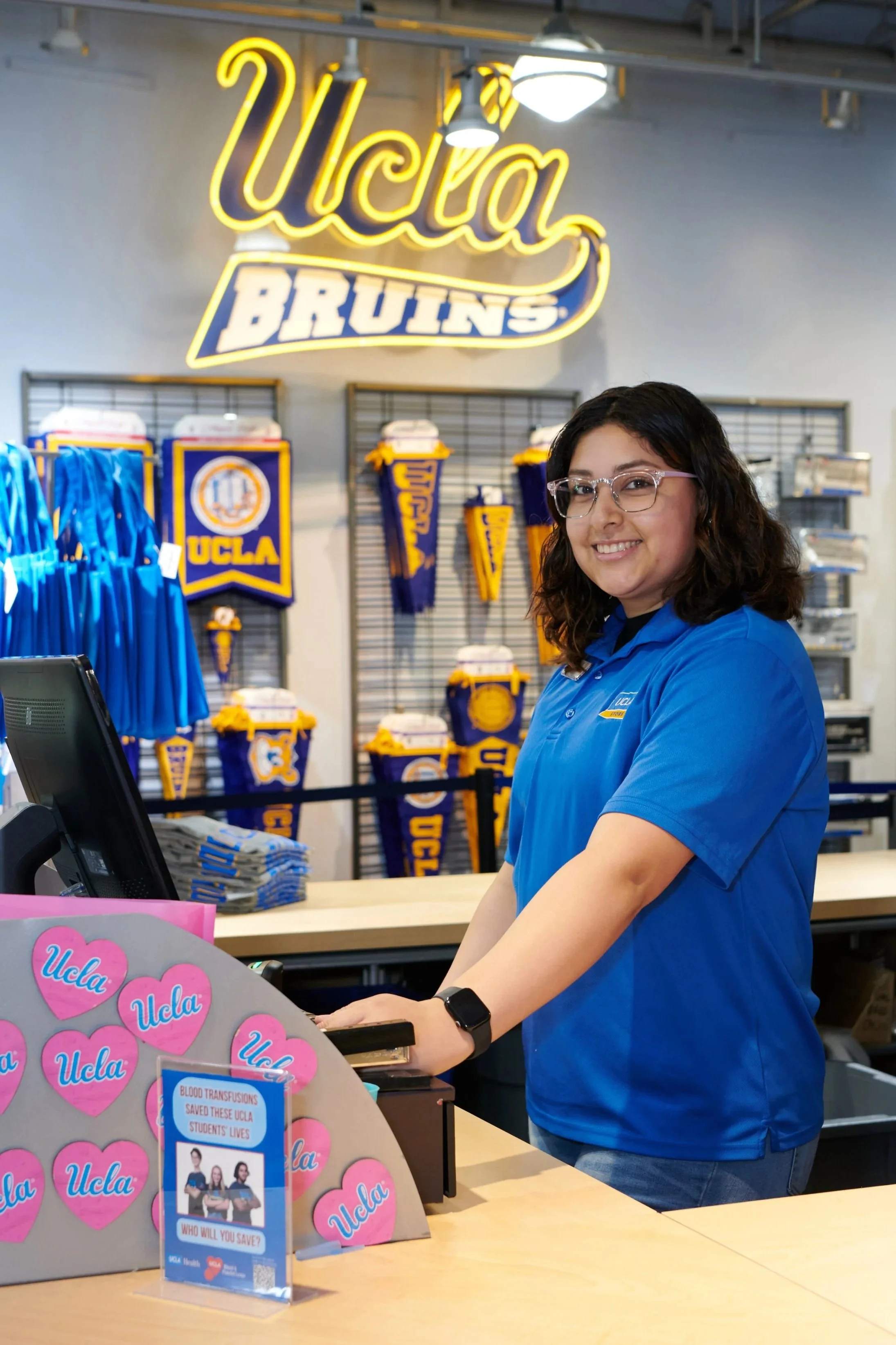 A smiling woman in a blue UCLA Store polo standing behind a cash register in front of a UCLA Bruins sign..