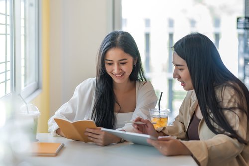 Two students studying together at a table, reviewing notes on a tablet and notebook with drinks beside them